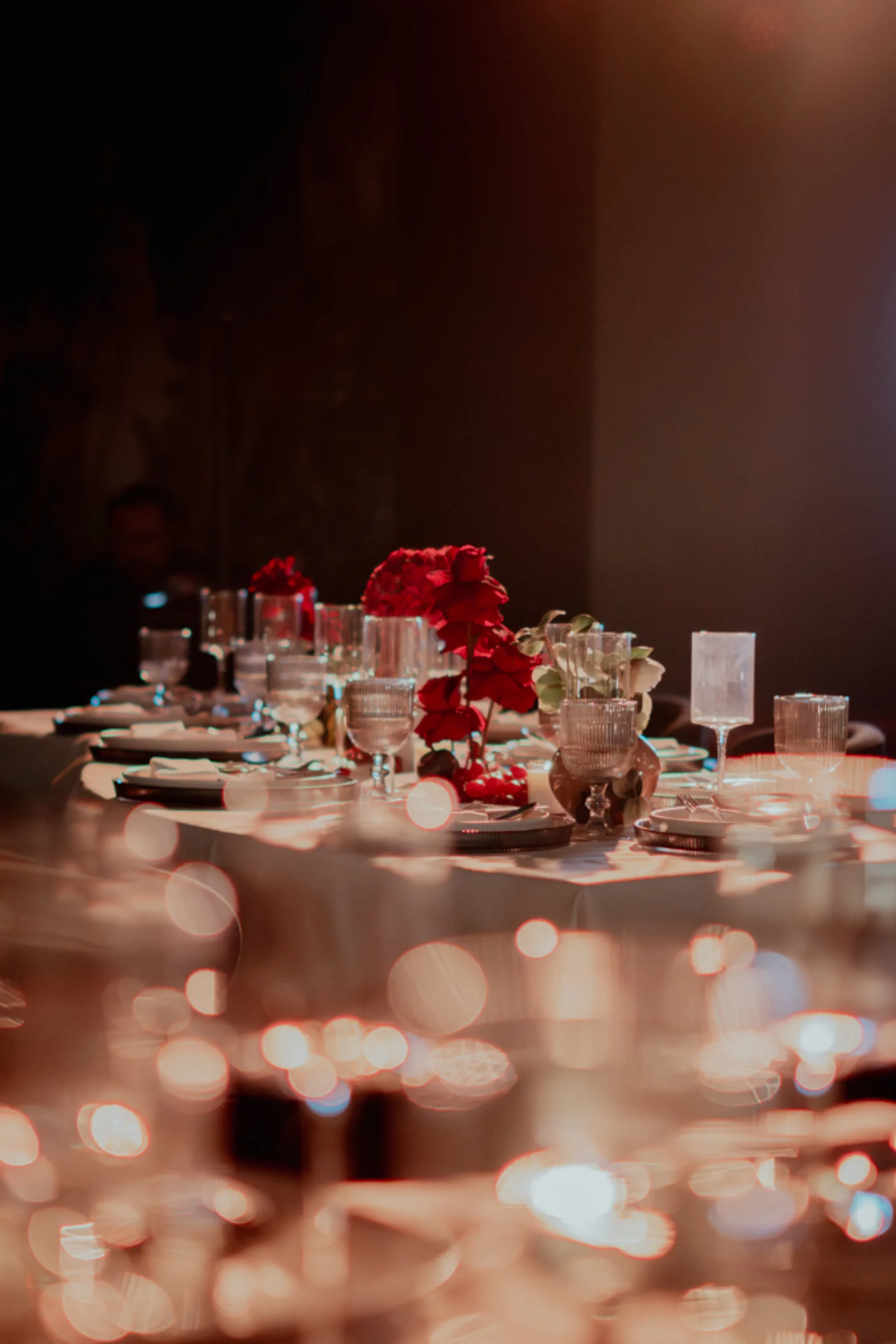 Mesa de comedor elegantemente dispuesta con cristalería brillante, flores rojas como pieza central, iluminación suave que crea un ambiente cálido y acogedor y un fondo borroso.