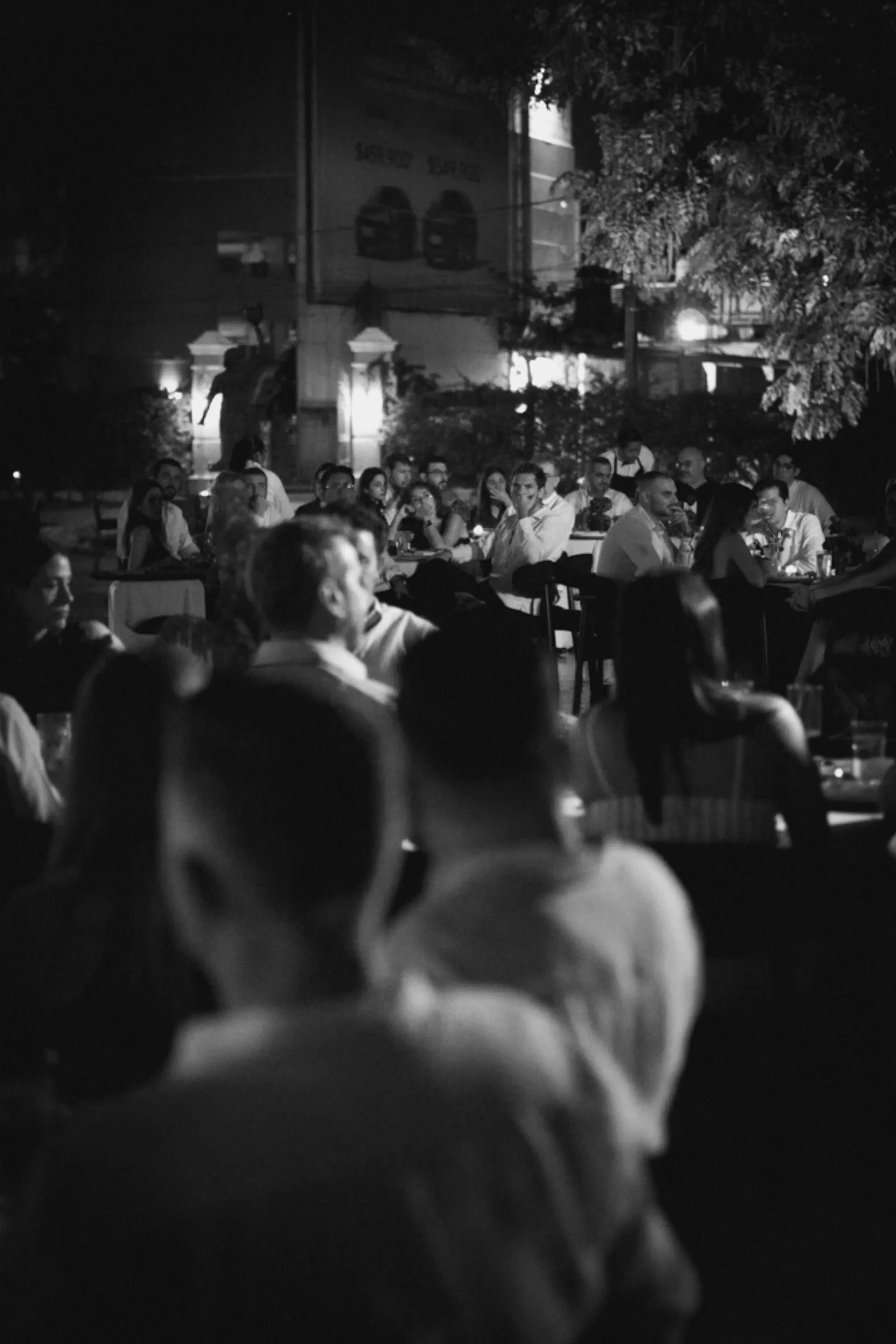 Una reunión al aire libre, en blanco y negro y con poca luz, con gente sentada en mesas. El ambiente es animado y social, con conversaciones y risas.