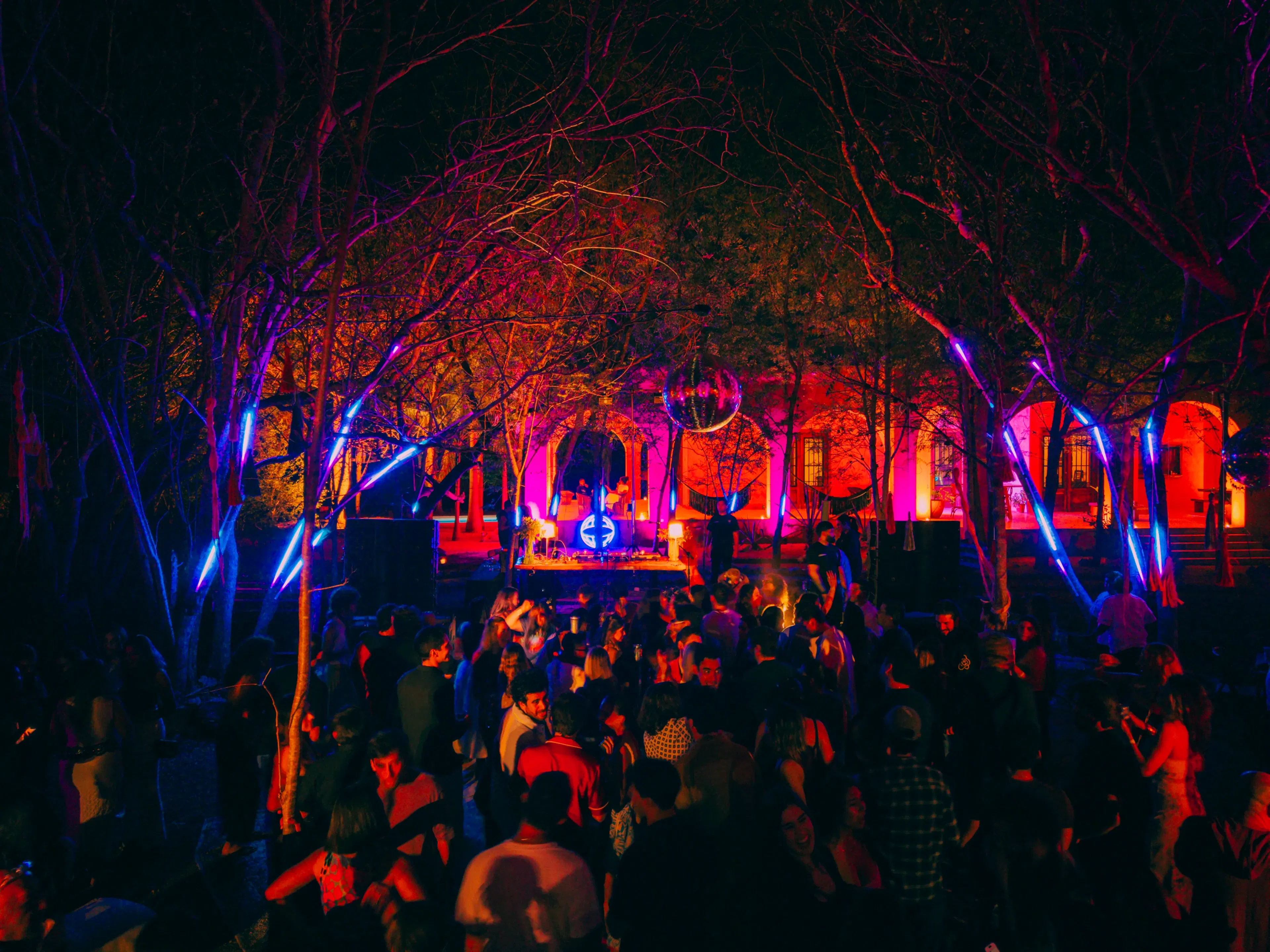 Una animada fiesta nocturna al aire libre con iluminación colorida. Una cabina de DJ, situada entre los árboles, ilumina al público con vibrantes luces rojas y azules, creando un ambiente vibrante.