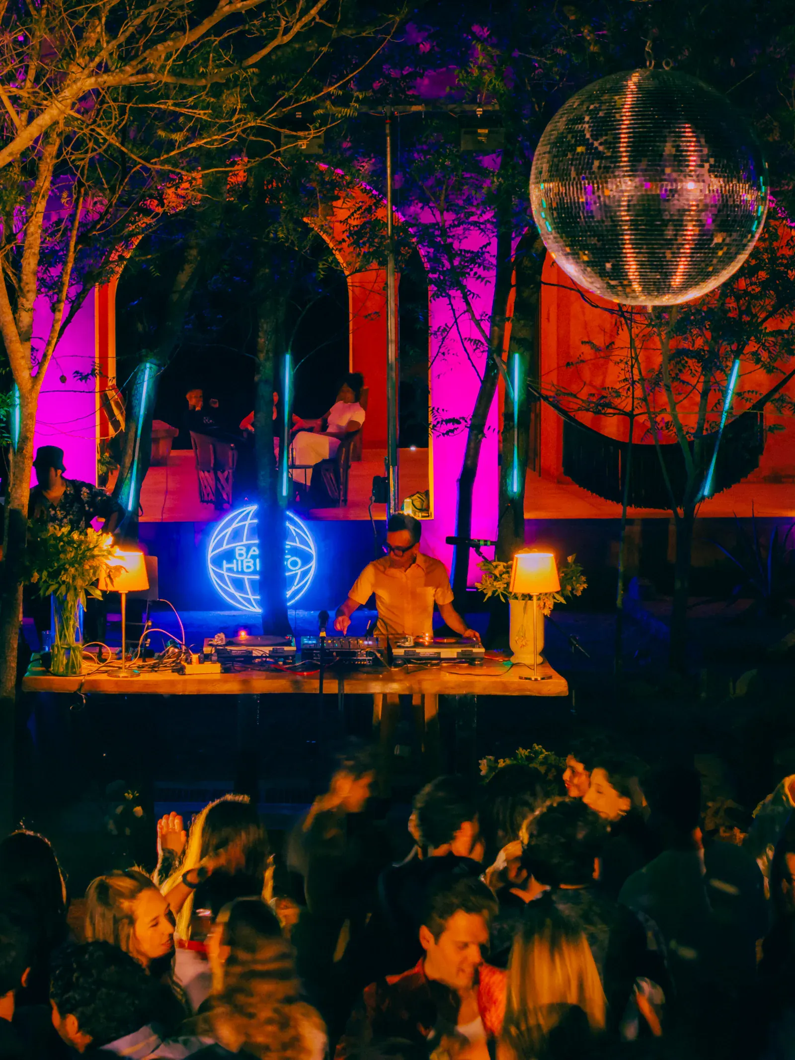 Un DJ pincha música en una fiesta al aire libre, rodeado de una multitud animada. Luces de neón brillan de fondo bajo una gran bola de discoteca, creando un ambiente festivo.