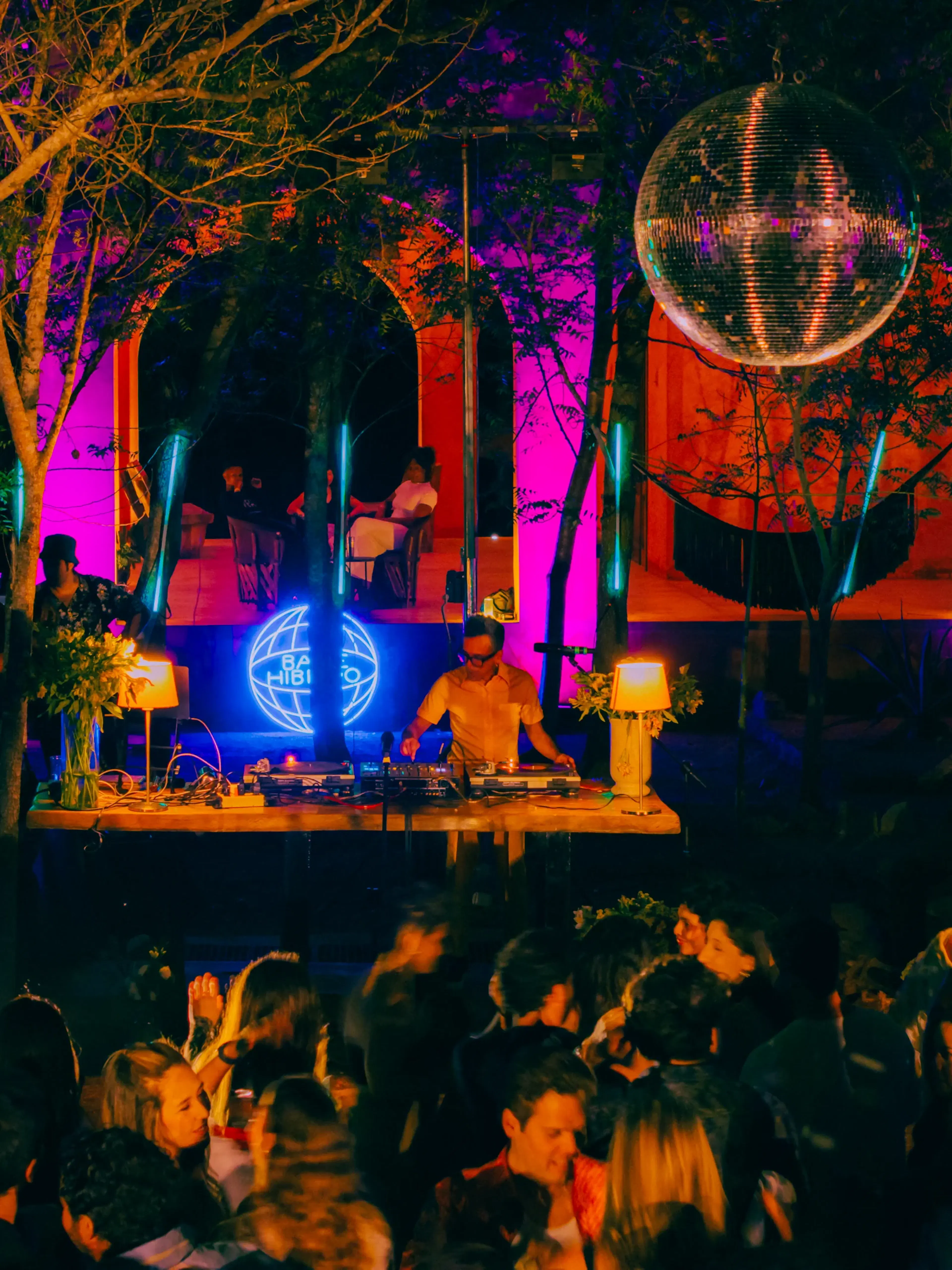 Un DJ pincha música en una fiesta al aire libre, rodeado de una multitud animada. Luces de neón brillan de fondo bajo una gran bola de discoteca, creando un ambiente festivo.