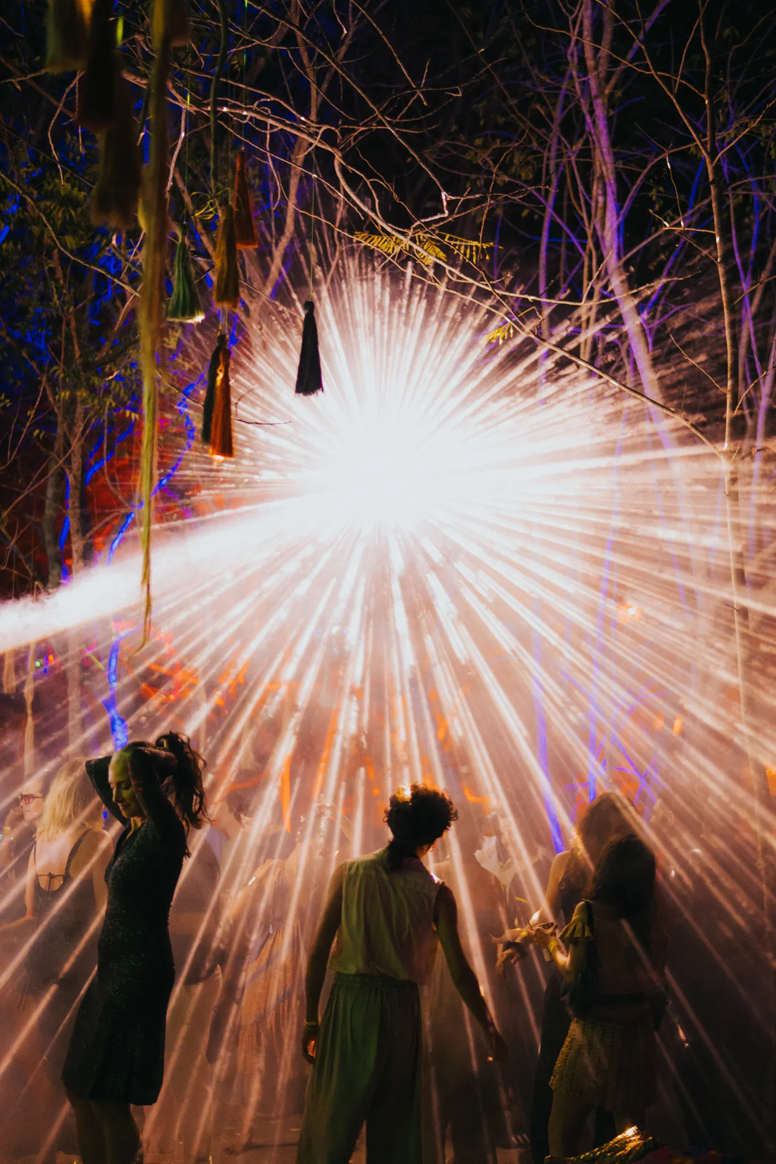 La gente baila bajo luces radiales brillantes que crean un deslumbrante efecto de estrella en un entorno boscoso al aire libre, evocando una atmósfera vibrante y festiva.