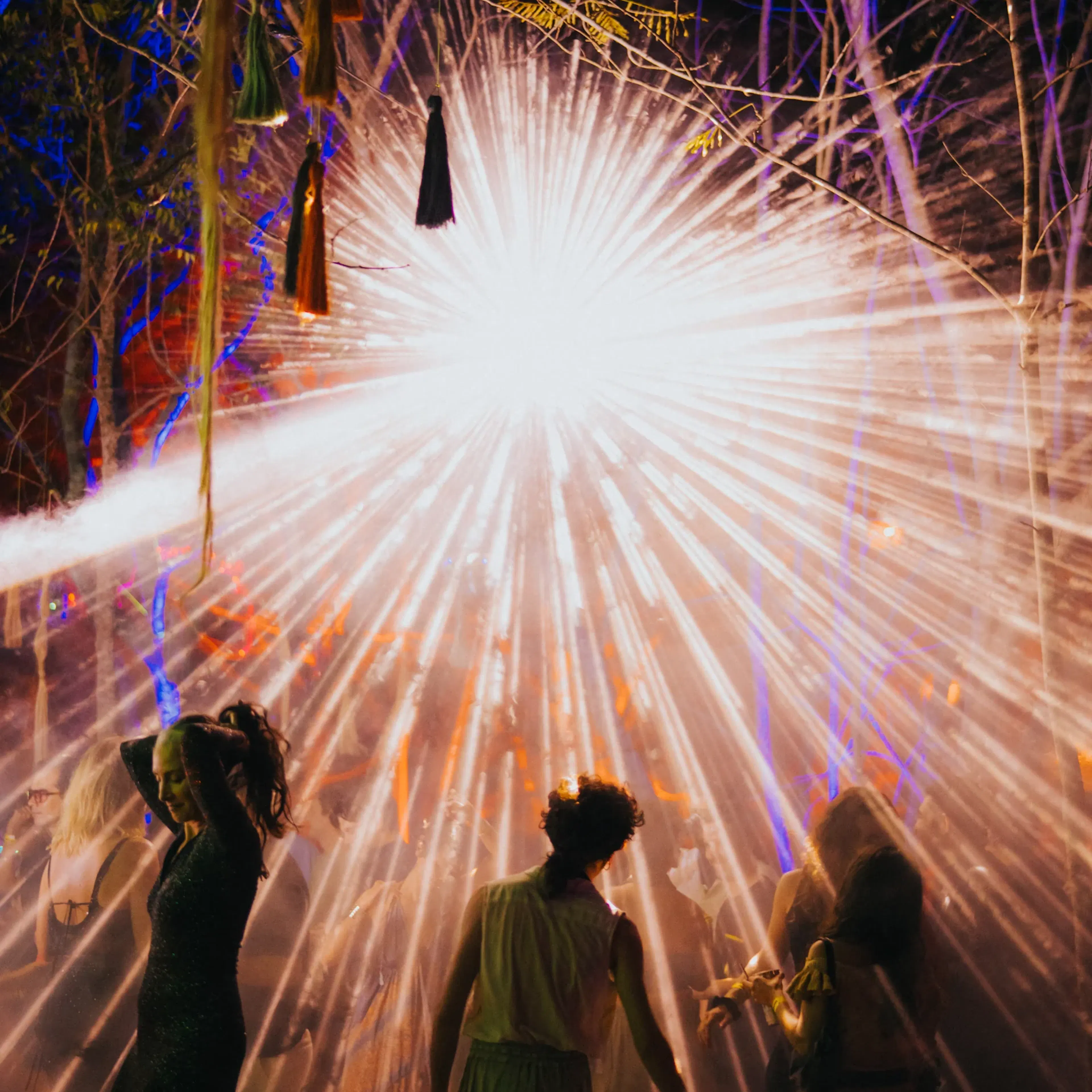 La gente baila bajo luces radiales brillantes que crean un deslumbrante efecto de estrella en un entorno boscoso al aire libre, evocando una atmósfera vibrante y festiva.