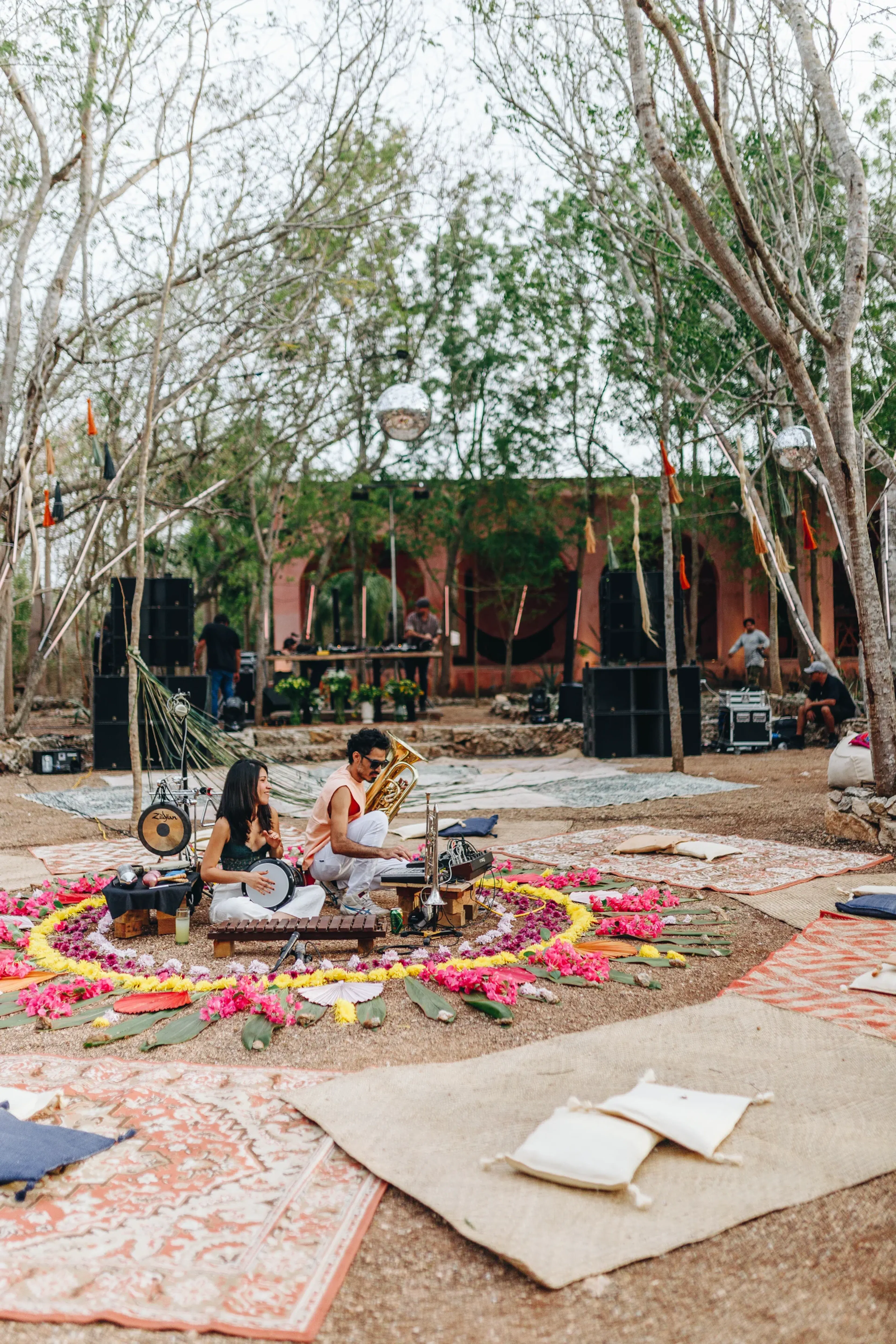 Dos músicos tocan instrumentos en un vibrante entorno al aire libre, adornado con flores y alfombras de colores. Altos árboles los rodean, creando un ambiente sereno y artístico.