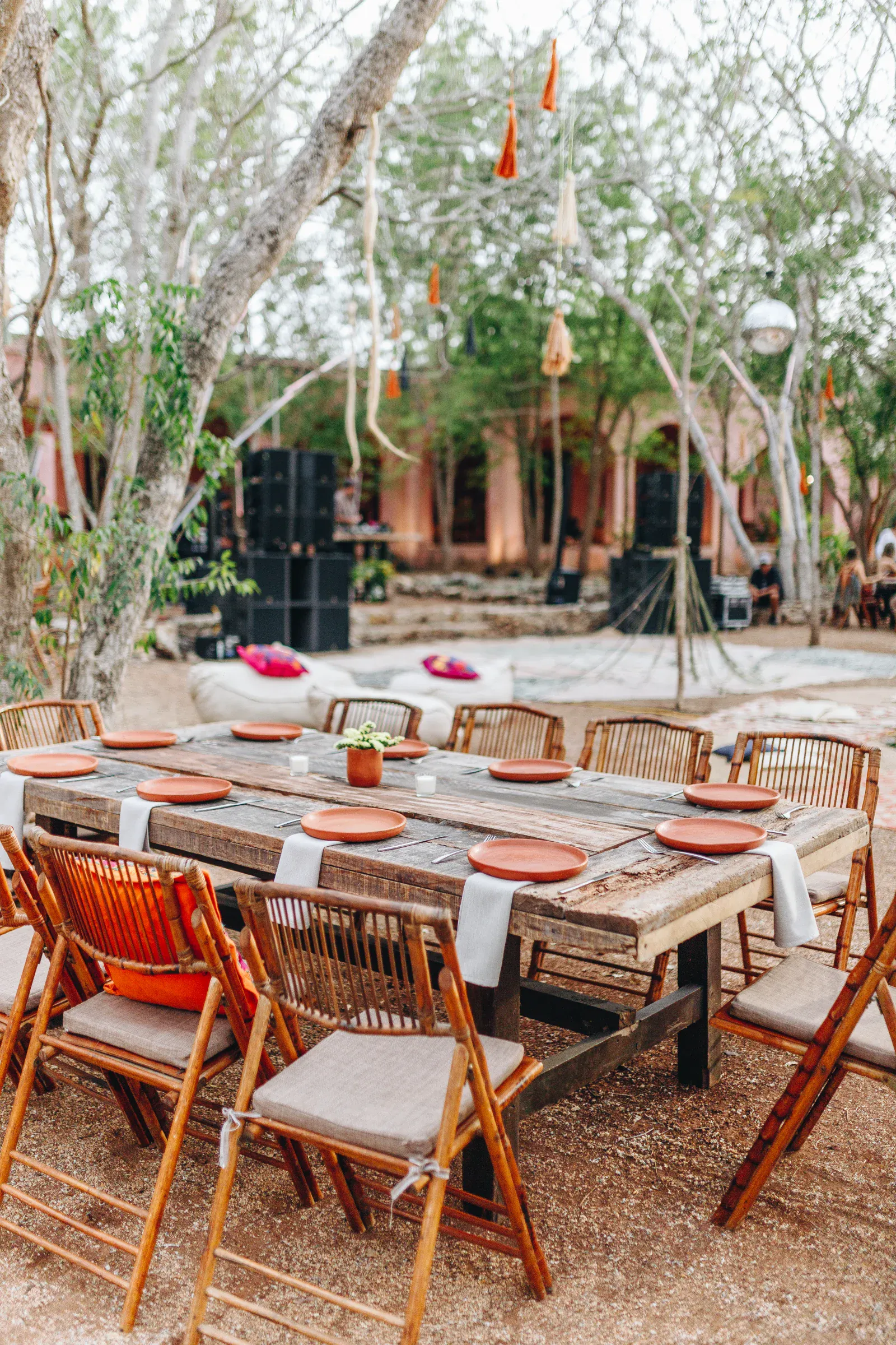 Comedor al aire libre con mesa rústica de madera y sillas de mimbre para ocho personas. Platos de terracota y servilletas blancas adornan la mesa, rodeada de árboles.