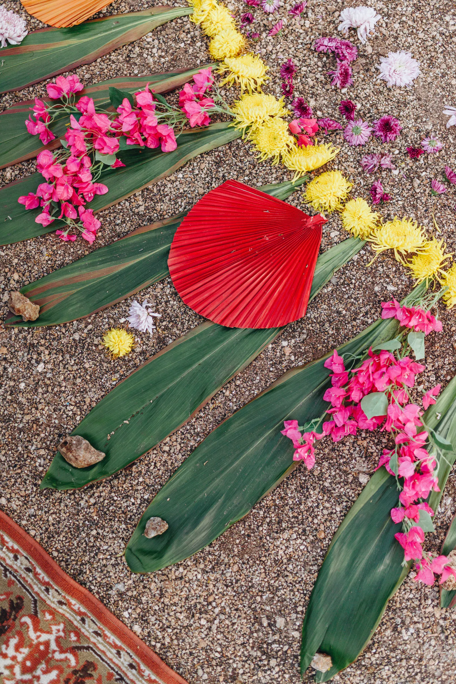 El colorido arreglo floral sobre grava presenta grandes hojas verdes, vibrantes flores rosas y amarillas, un abanico rojo y pequeñas rocas, creando una atmósfera festiva.