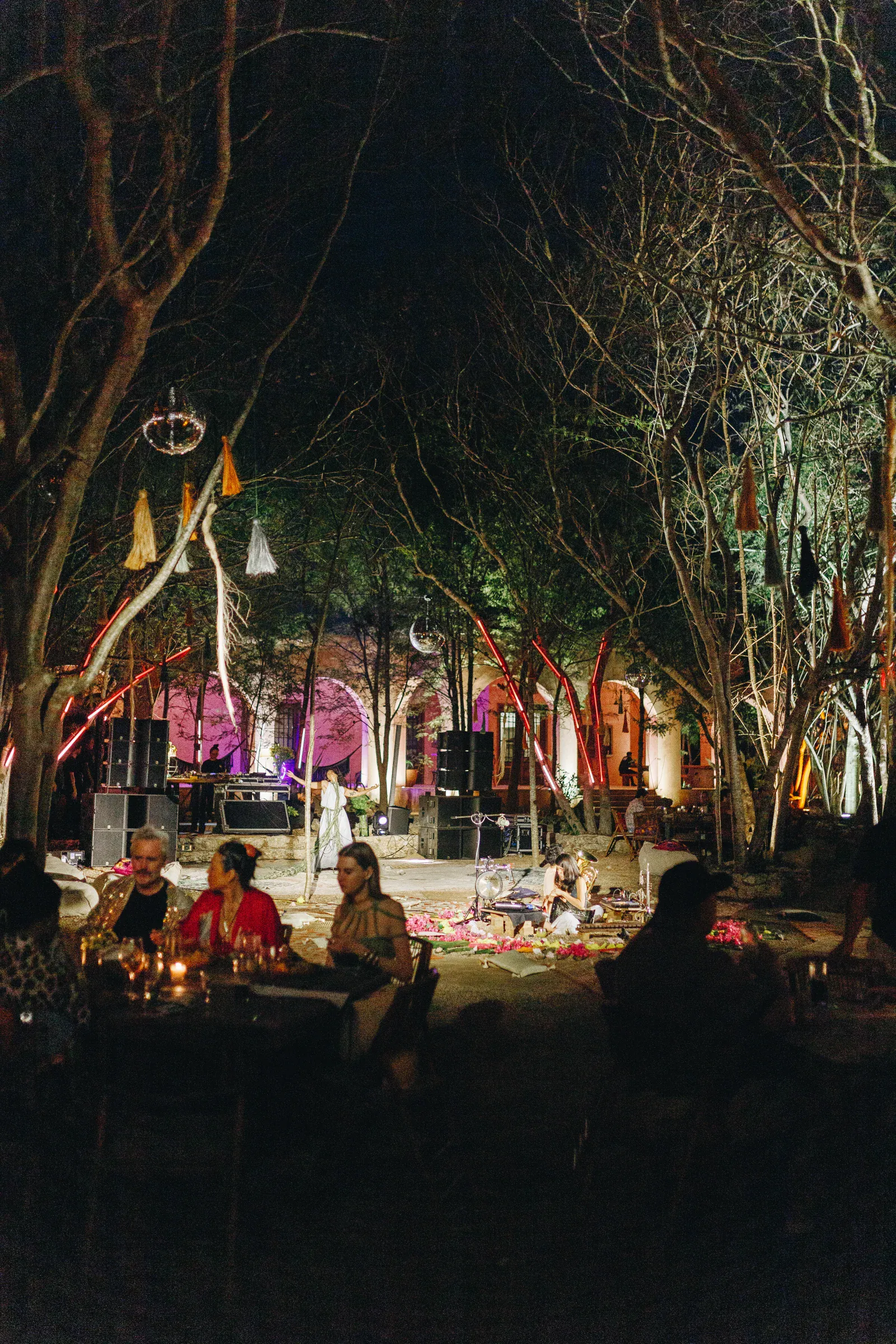 Reunión vespertina al aire libre bajo árboles desnudos y luces de colores. La gente se sienta en mesas y charla. Los altavoces y la decoración crean un ambiente festivo y ecléctico.