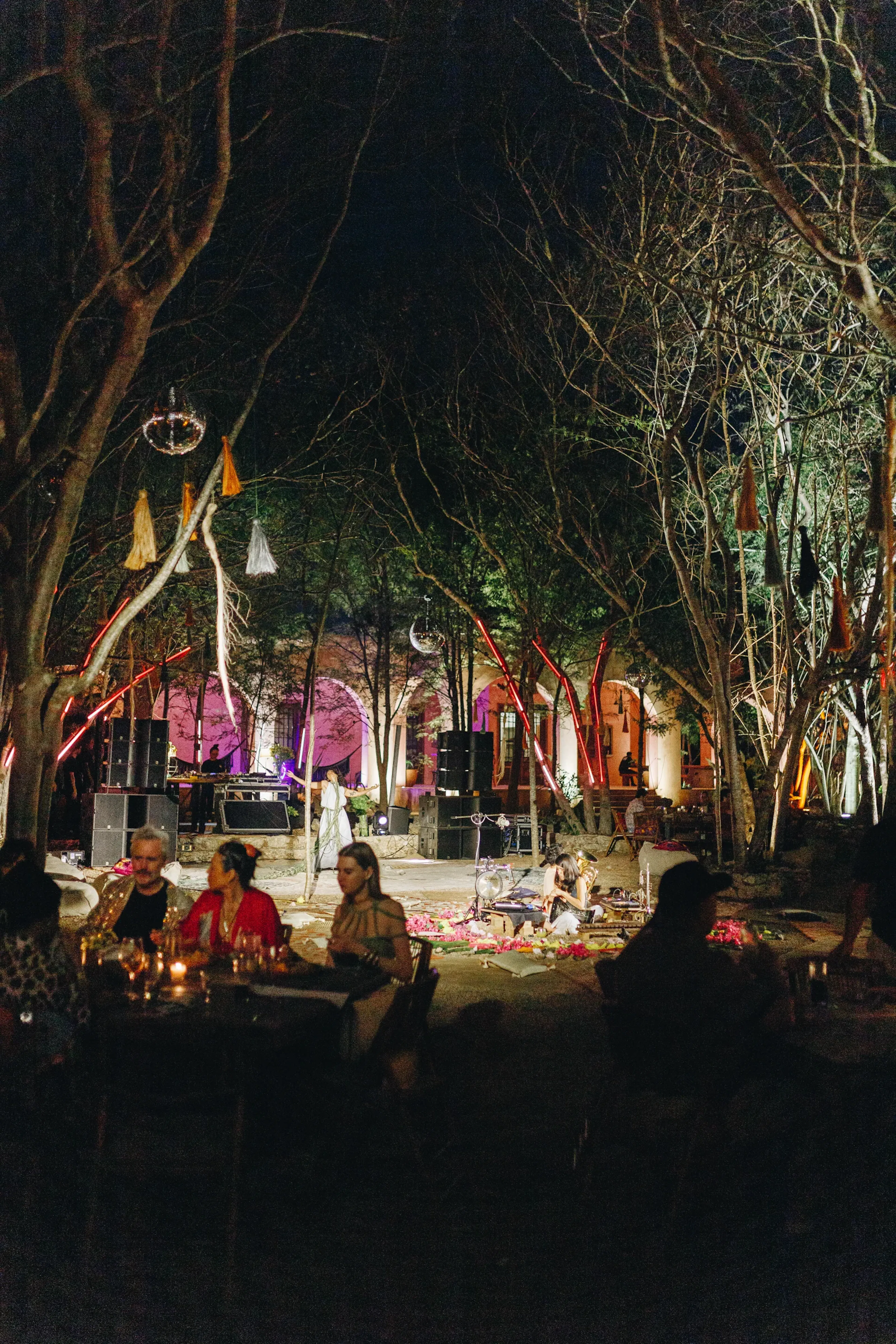 Reunión vespertina al aire libre bajo árboles desnudos y luces de colores. La gente se sienta en mesas y charla. Los altavoces y la decoración crean un ambiente festivo y ecléctico.