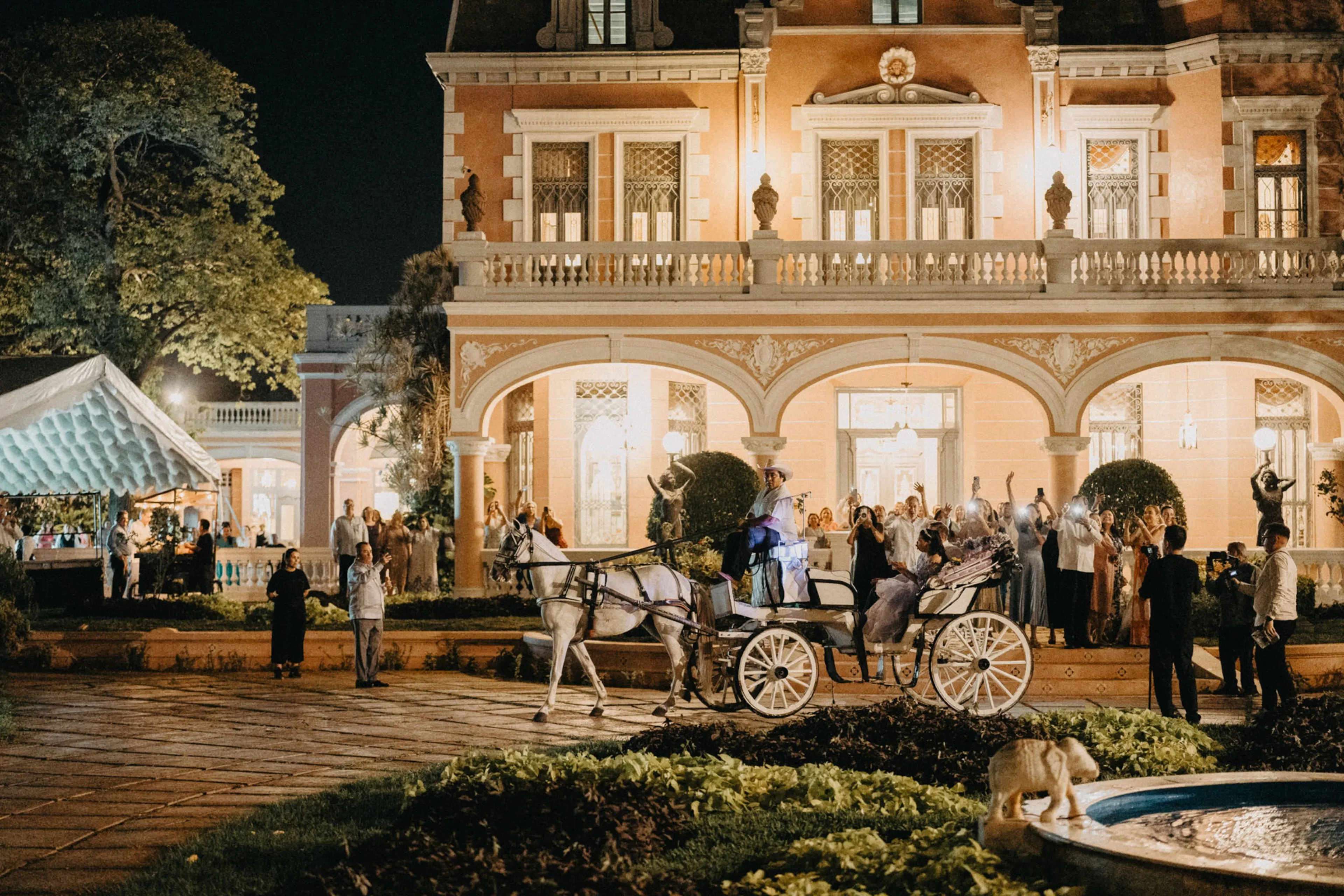 Una gran mansión iluminada sirve de telón de fondo para una noche festiva. Un carruaje tirado por caballos espera afuera, rodeado de gente elegantemente vestida y exuberante vegetación.