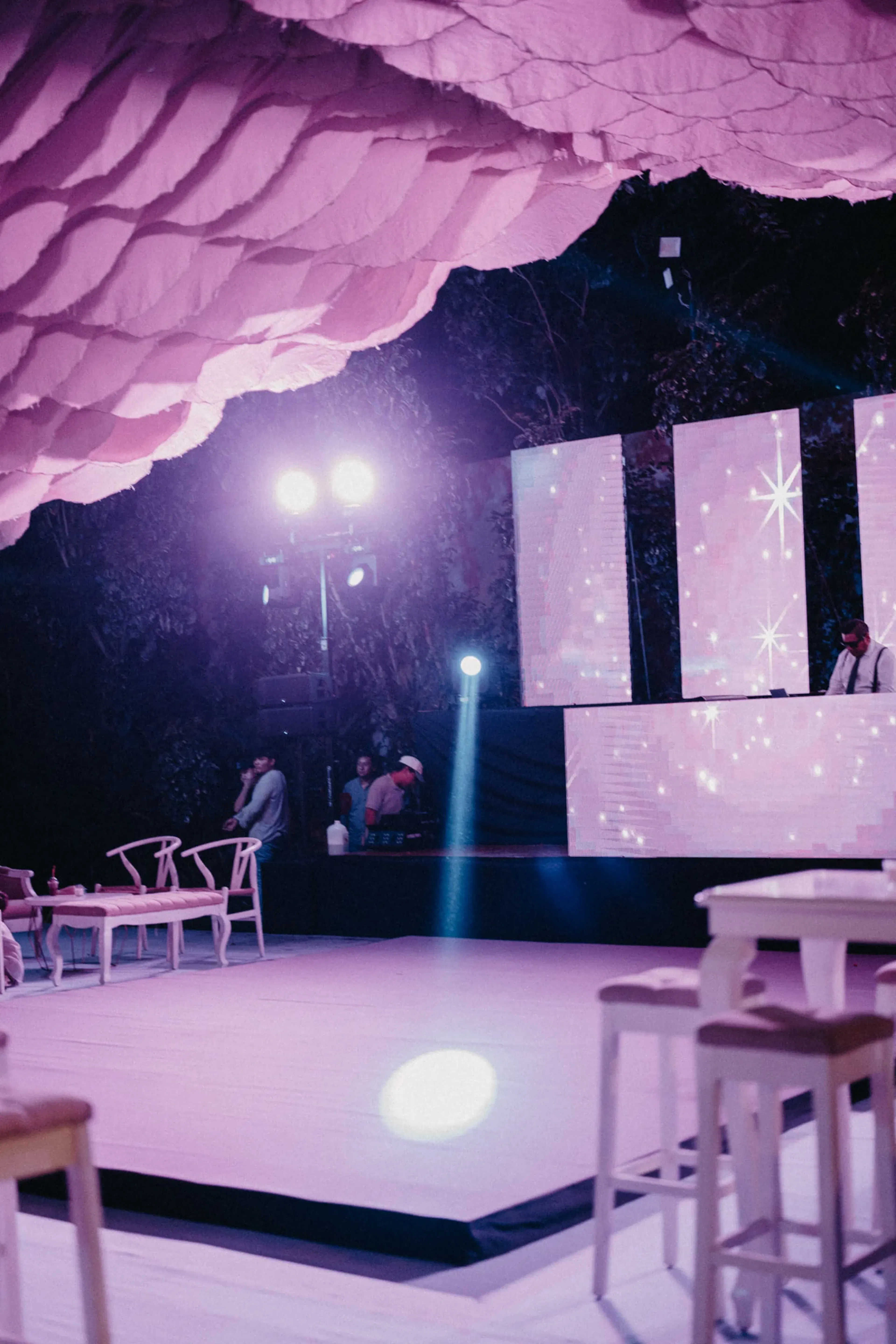 Un elegante espacio para eventos con iluminación rosa, techo de tela curva, un escenario iluminado con pantallas de estrellas y sillas y mesas vacías. El ambiente es moderno y elegante.