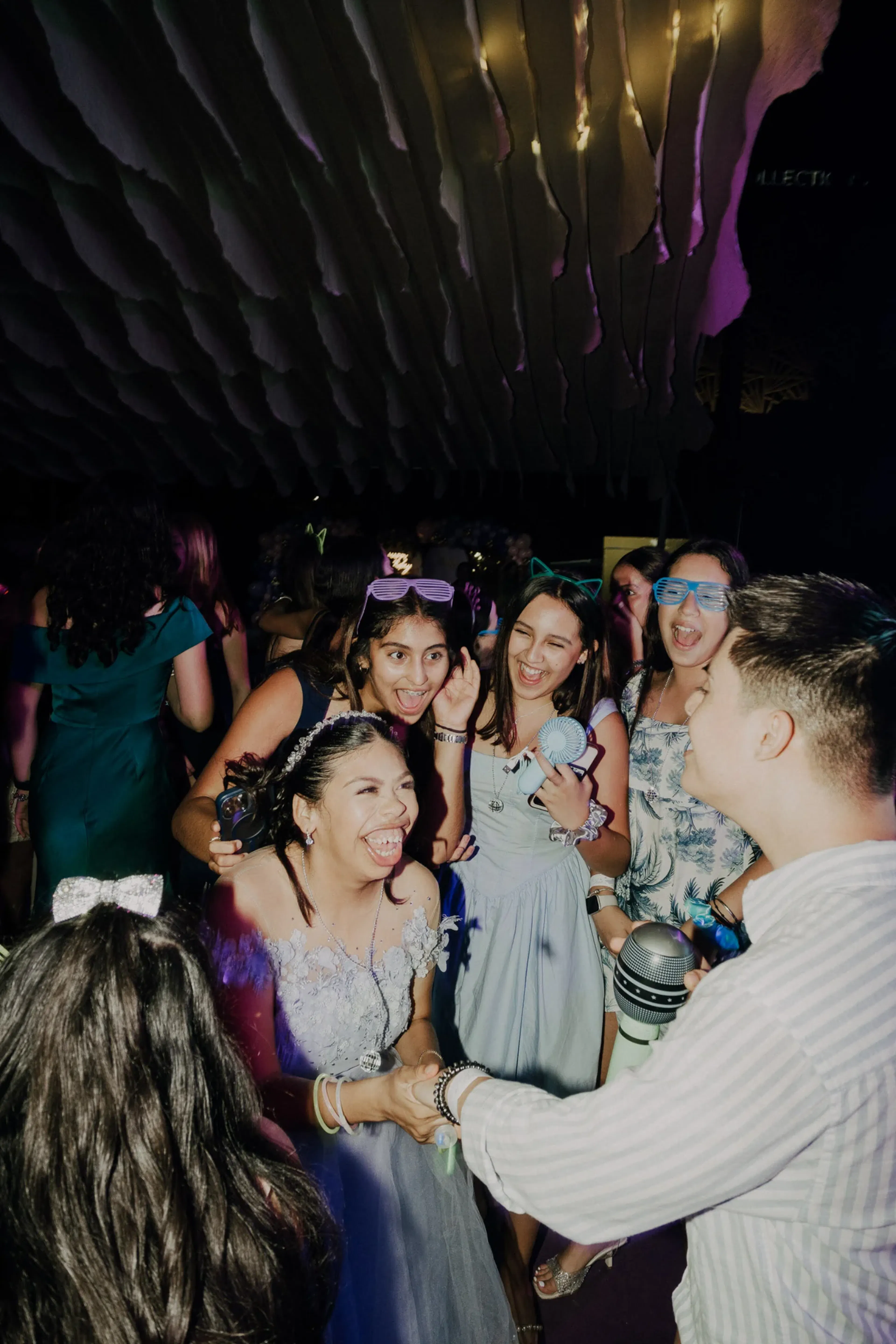 Un alegre grupo de jóvenes riendo y bailando en una fiesta, con luces y decoraciones coloridas. El ambiente es animado y festivo.