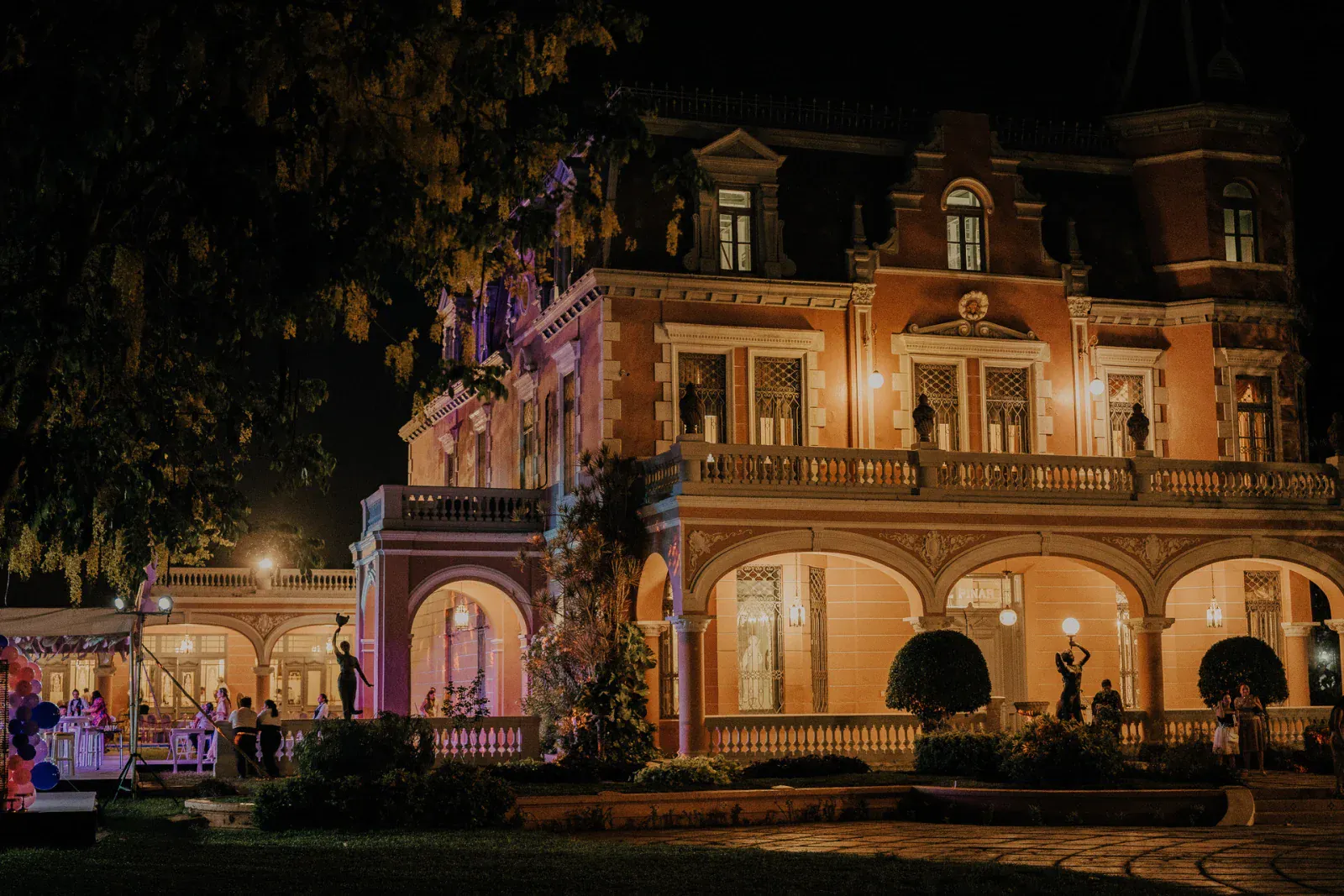 Iluminada por la noche, una gran mansión ornamentada brilla cálidamente con ventanas arqueadas y luces tenues. Los invitados se reúnen en la terraza, creando un ambiente elegante.