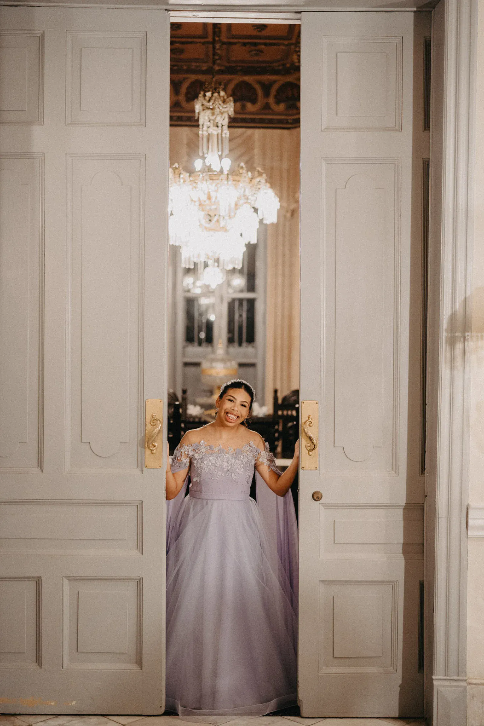 Una mujer con un elegante vestido lavanda sonríe cálidamente desde un imponente portal. Tras ella, brilla una lujosa lámpara de araña, añadiendo una sensación de opulencia.