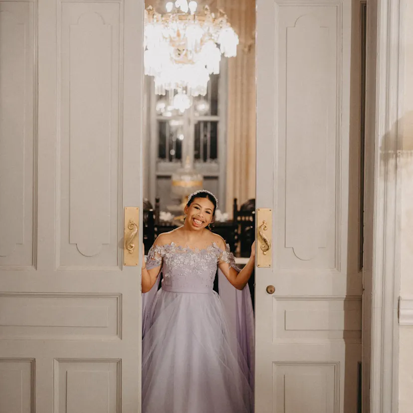 Una mujer con un elegante vestido lavanda sonríe cálidamente desde un imponente portal. Tras ella, brilla una lujosa lámpara de araña, añadiendo una sensación de opulencia.