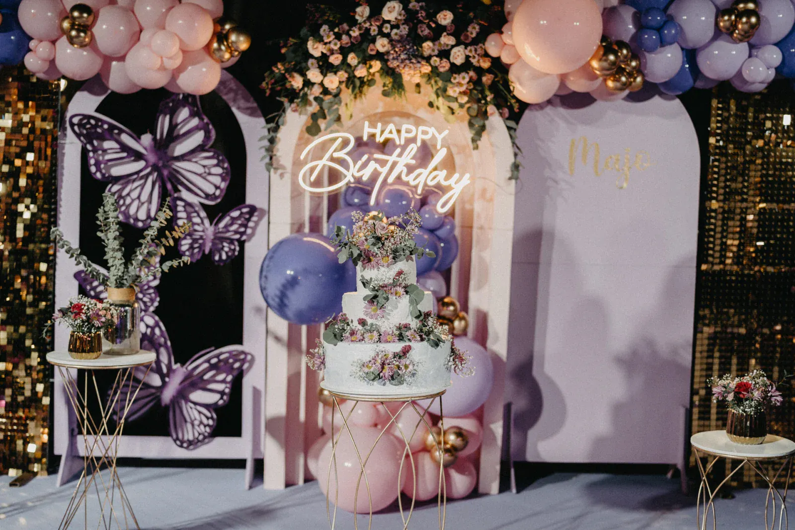 Elegante decoración de cumpleaños con un pastel floral sobre una base dorada. El letrero de "Feliz Cumpleaños" brilla en la parte superior, rodeado de globos rosas y morados, flores y un fondo de mariposas. Festivo y alegre.