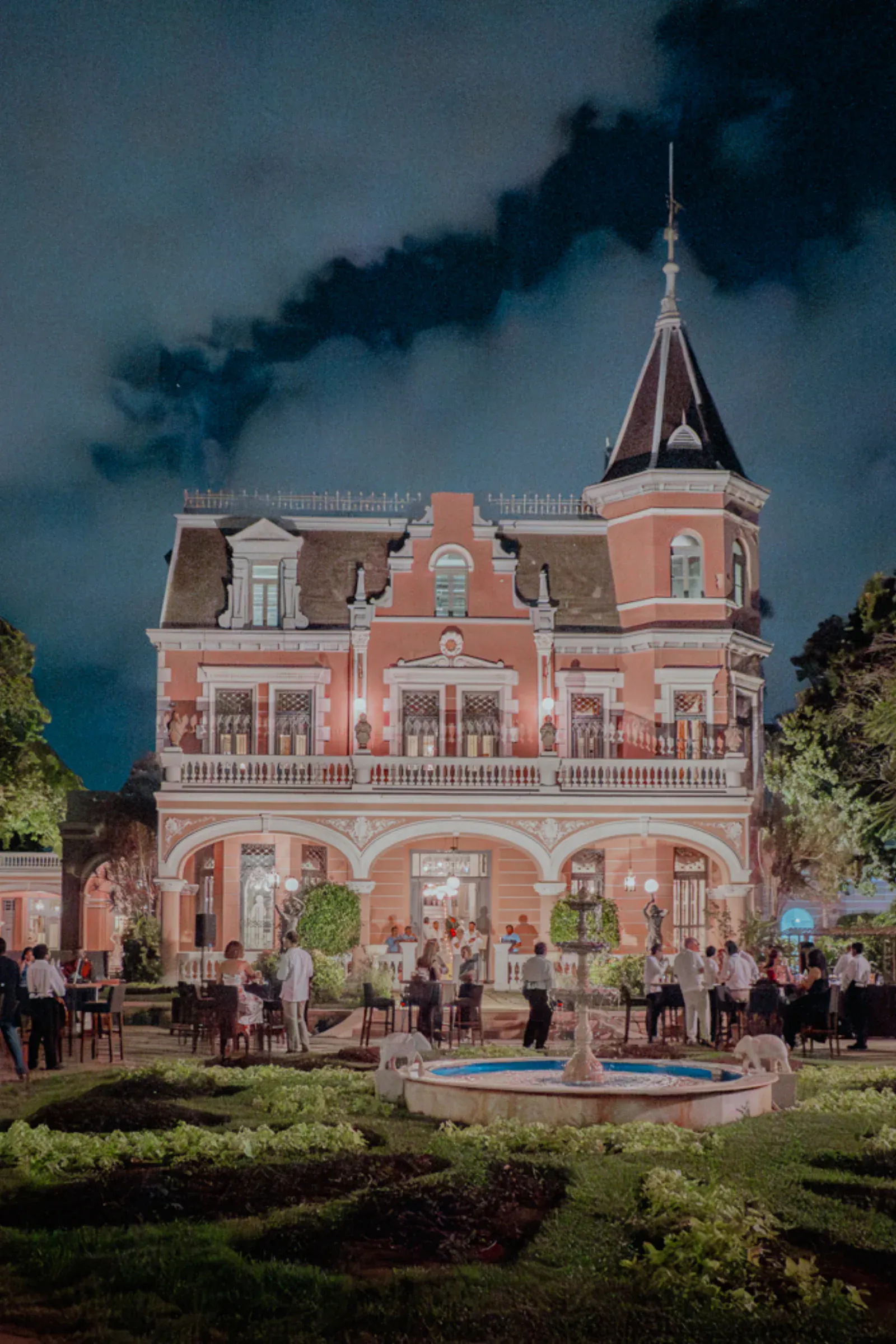Mansión roja ornamentada, iluminada por la noche, con una torre y ventanas arqueadas. Los invitados, vestidos de gala, socializan frente a la casa, cerca de una fuente iluminada.
