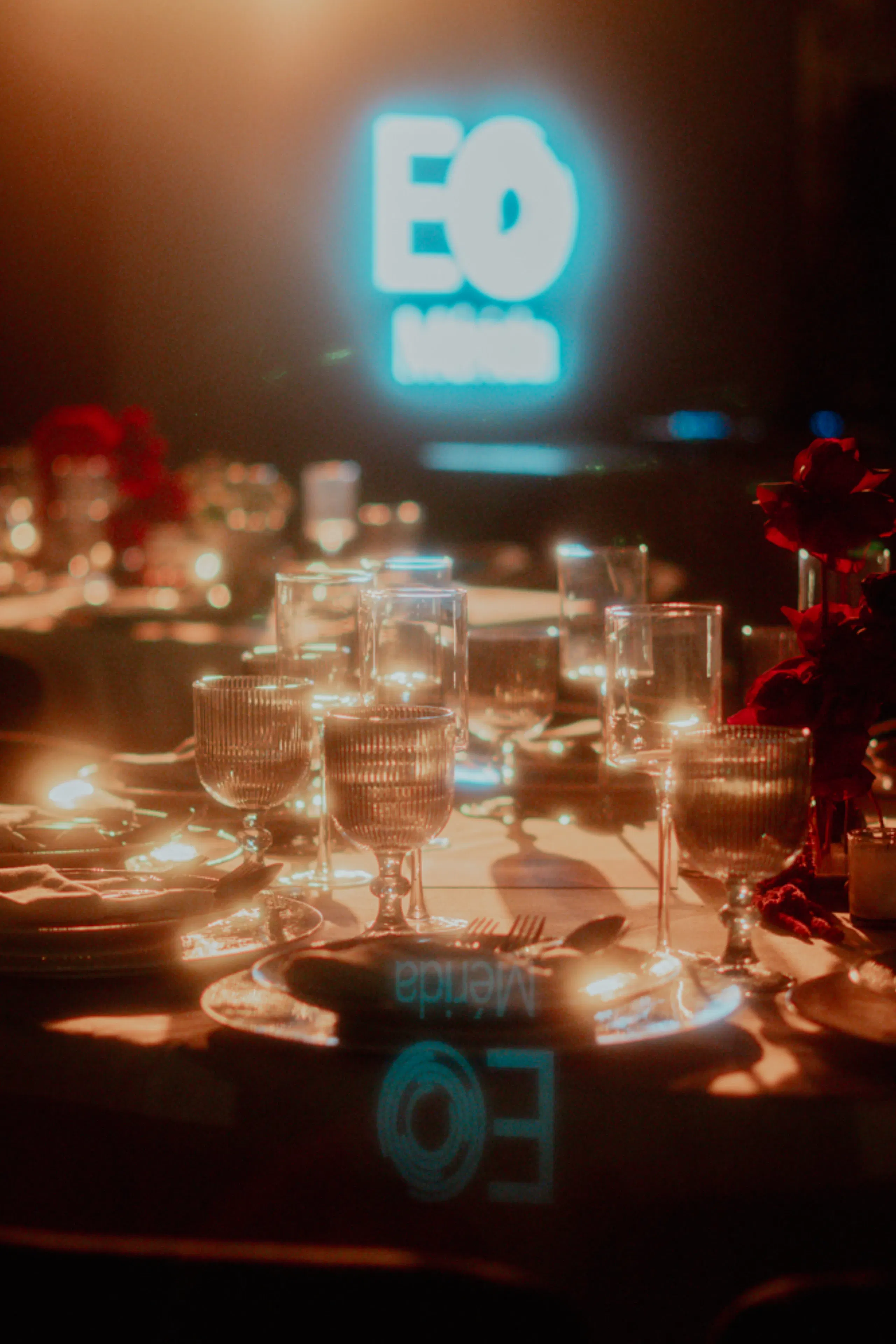 Elegante mesa con velas encendidas, cristalería y flores rojas. Un letrero de neón azul de fondo le da un toque moderno.