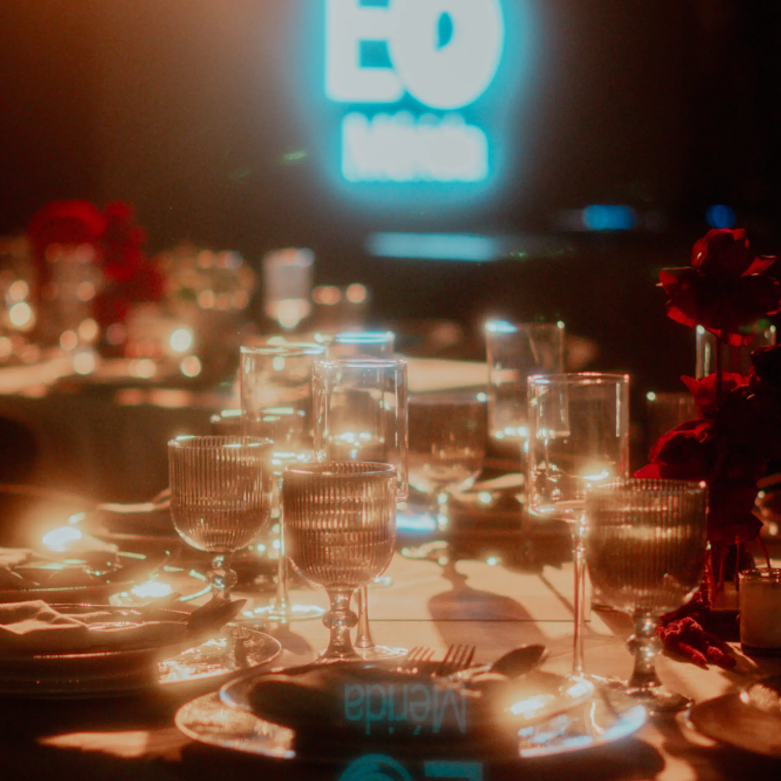 Elegante mesa con velas encendidas, cristalería y flores rojas. Un letrero de neón azul de fondo le da un toque moderno.