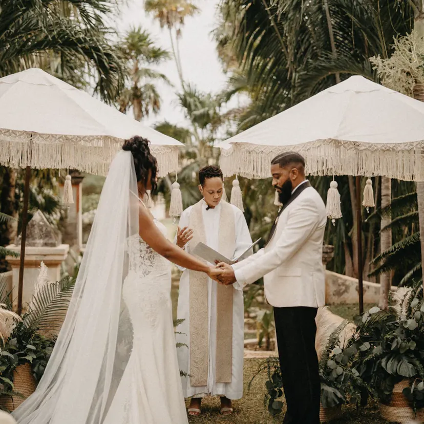 Los novios se toman de la mano bajo sombrillas con flecos, con un celebrante en el centro. Una exuberante vegetación los rodea, creando un elegante ambiente tropical para la boda.