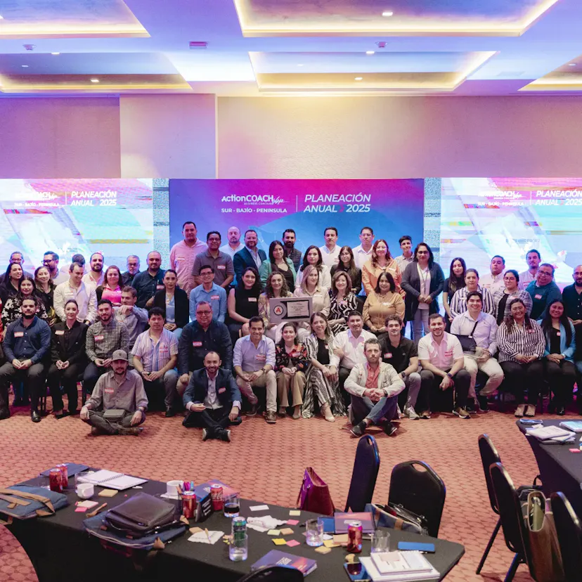 Un grupo numeroso de profesionales posa en una sala de conferencias para una foto de equipo. Al fondo se ve una pantalla digital con el texto "Planeación Anual 2025". El ambiente es profesional y colaborativo. En primer plano se ven mesas con papeles y bebidas.