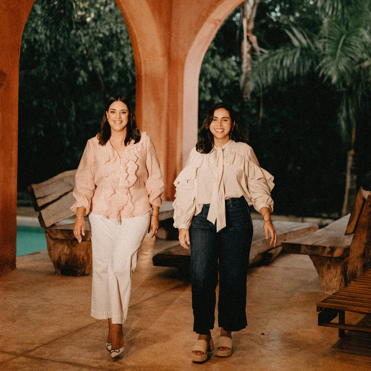 Dos mujeres con blusas con volados caminan en un patio de tonos cálidos, con vegetación tropical visible a través de puertas arqueadas, creando una atmósfera relajada, amigable y profesional.