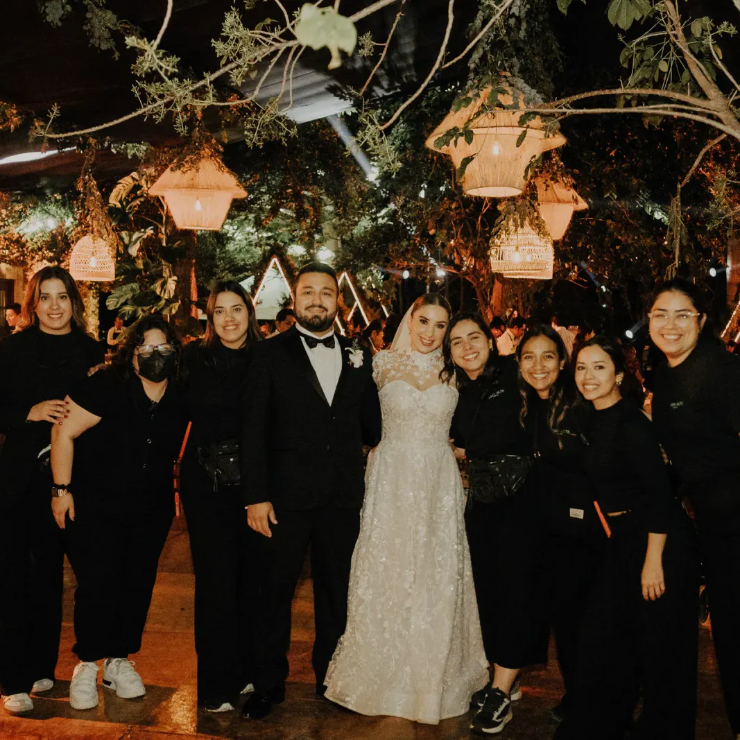 Una foto grupal en una boda muestra a una novia con vestido blanco y a un novio con esmoquin, rodeados de gente sonriente vestida de negro. Iluminación cálida y festiva.