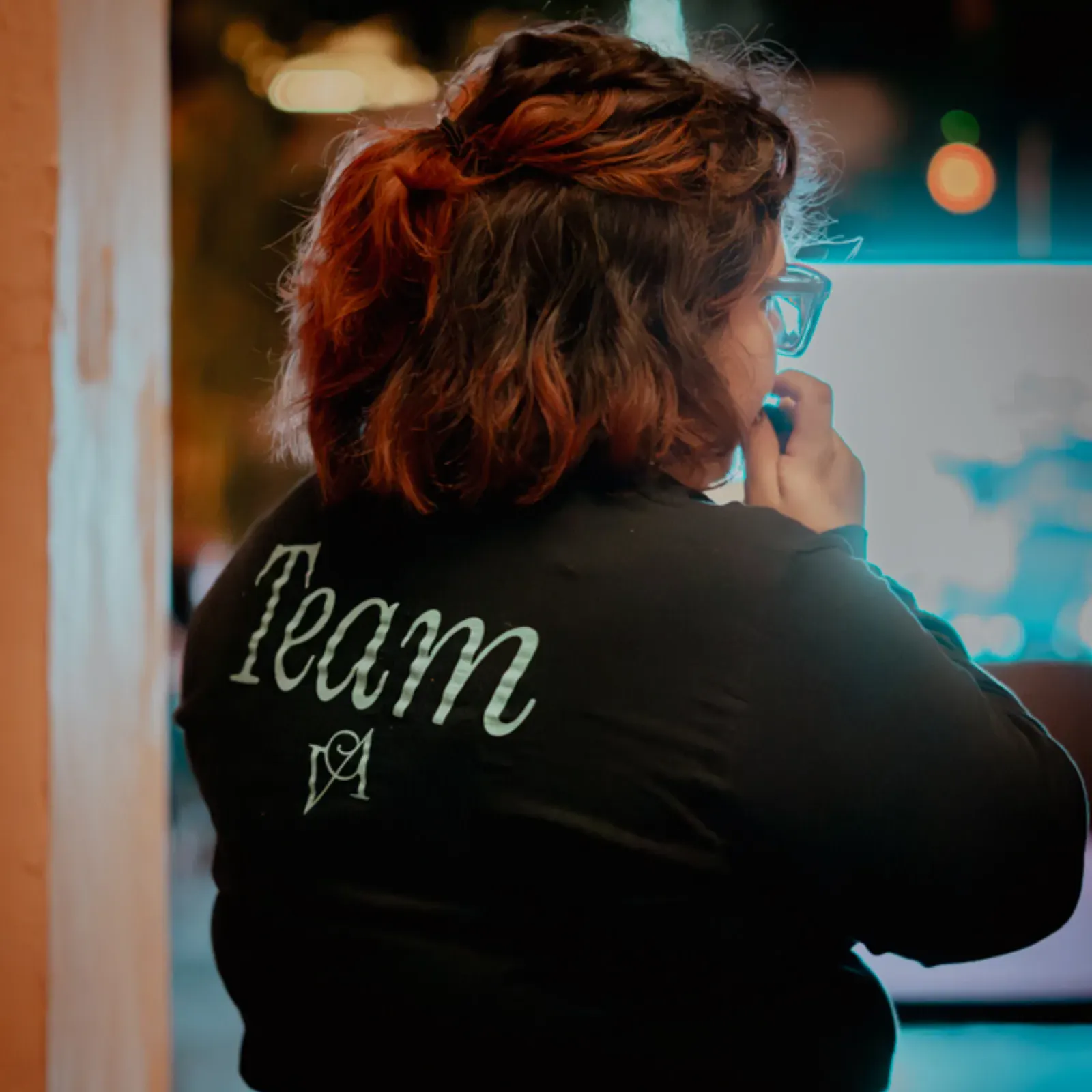 Una persona con el pelo hasta los hombros está de pie en un interior, de espaldas. Lleva una chaqueta con la palabra "Team Vara Alta" en la espalda y parece pensativa. Iluminación cálida y suave.