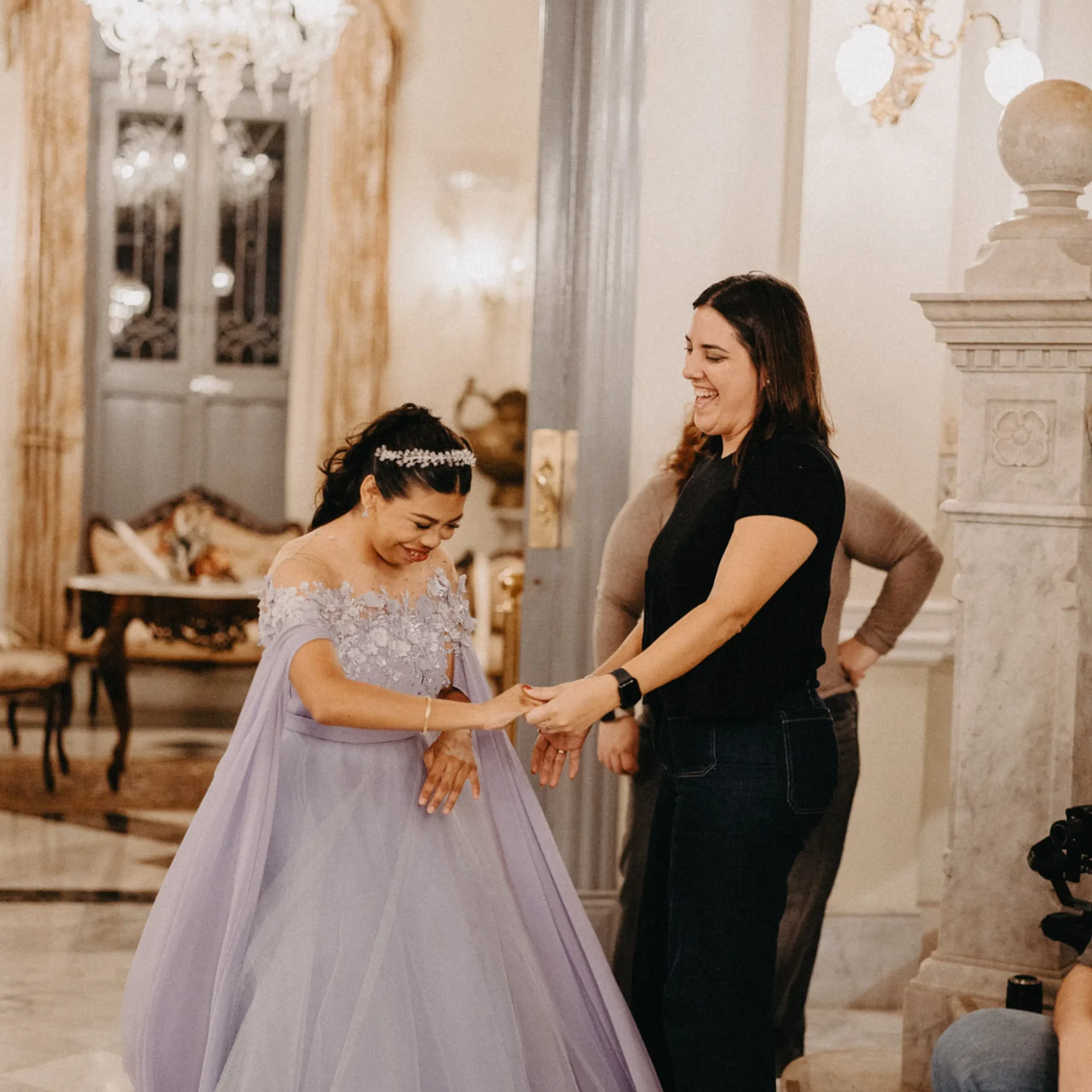 Una mujer sonriente con un elegante vestido de color violeta claro se encuentra en una gran habitación con una decoración ornamentada y candelabros, interactuando alegremente con otra mujer.