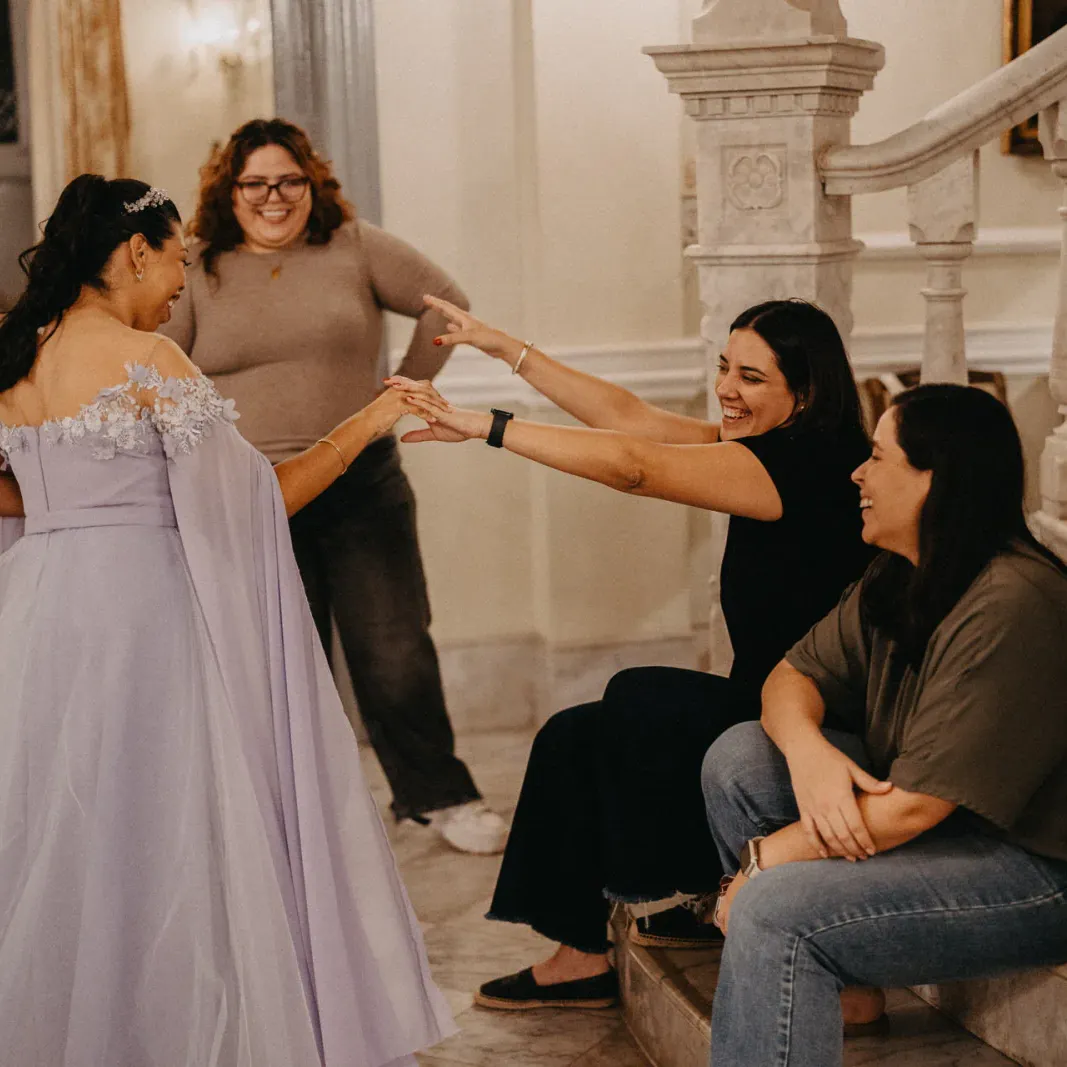 Una mujer alegre con un vestido lavanda se acerca a tres amigas sentadas en una escalera de mármol. Sonríen, creando un ambiente cálido y festivo.