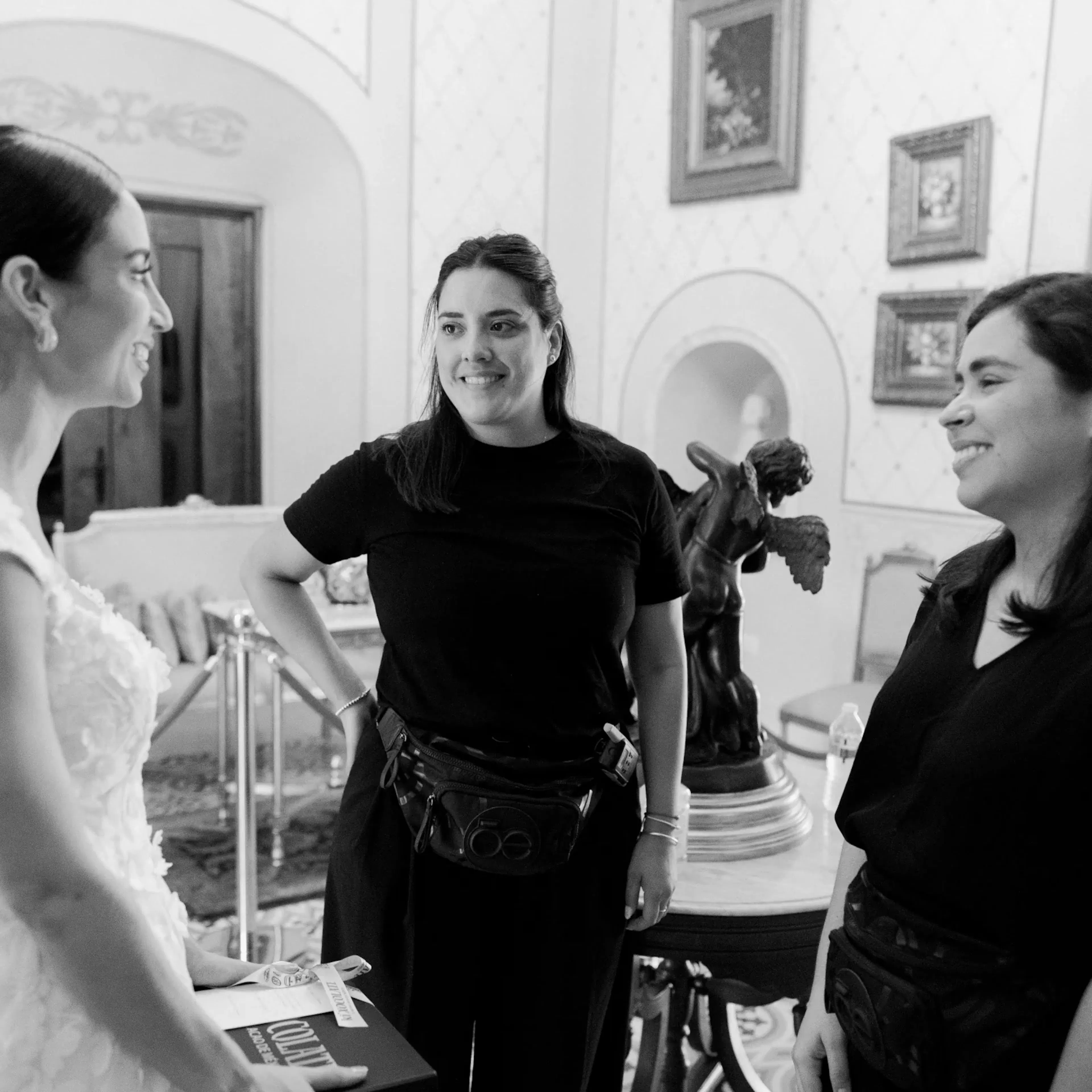 Una novia alegre con un vestido blanco interactúa con dos mujeres sonrientes con trajes negros dentro de una habitación ornamentada con arte en las paredes y una escultura sobre una mesa.