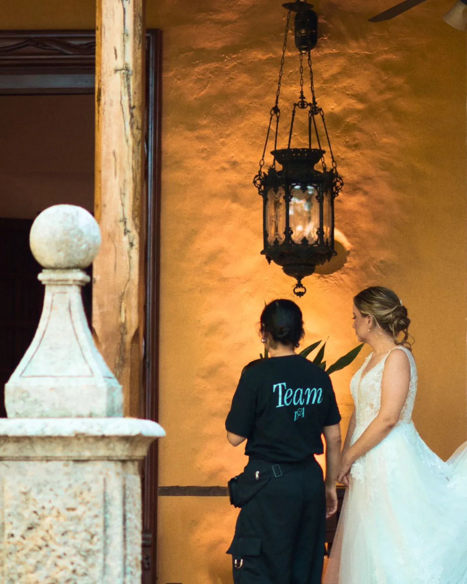 Una novia con un vestido blanco está de pie junto a un miembro del equipo vestido de negro, bajo una linterna rústica contra una pared amarilla cálida y texturizada, creando una atmósfera serena.