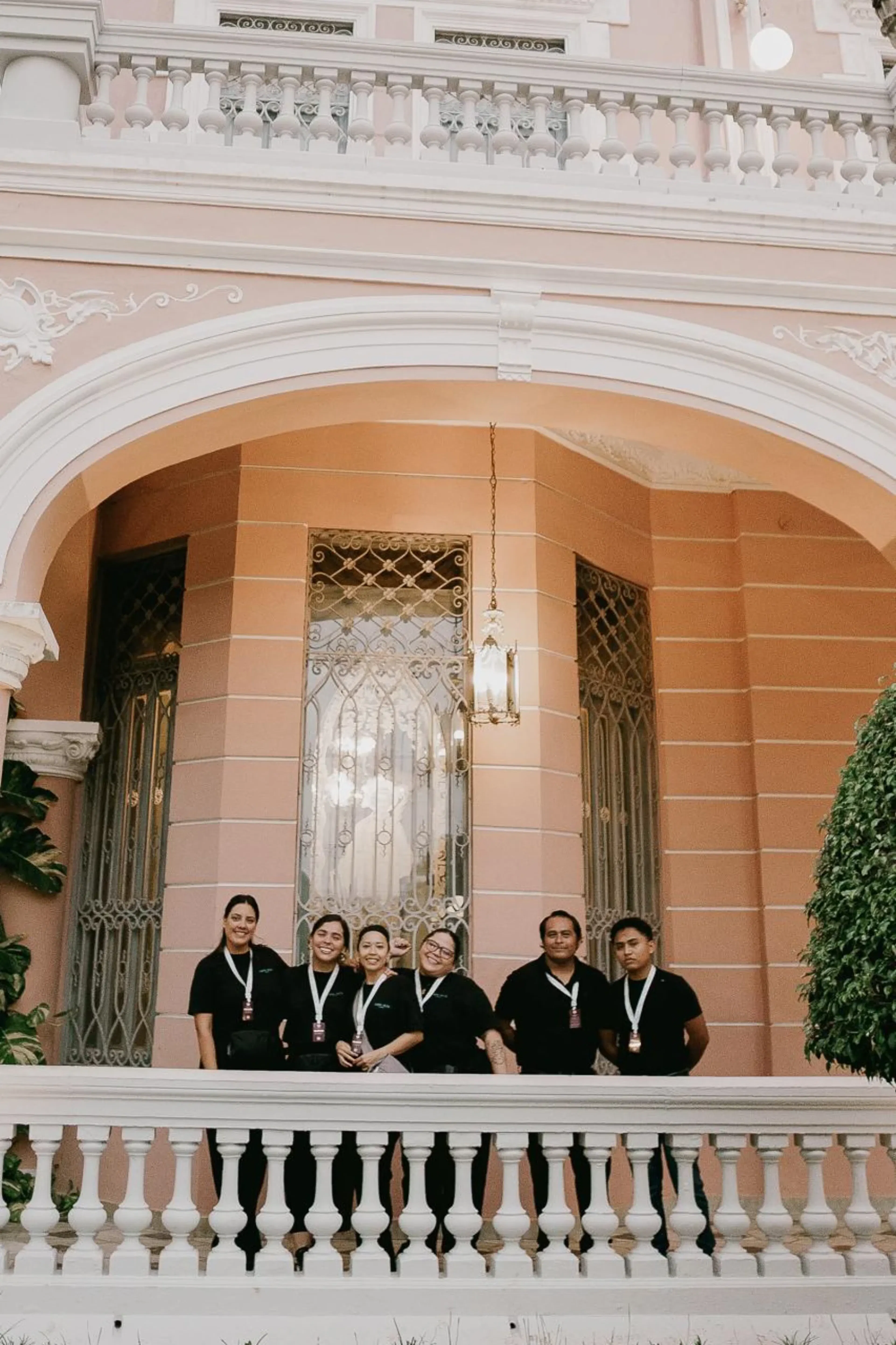 Un grupo de seis personas vestidas de negro se encuentran sonriendo frente a un edificio de estuco rosa con herrería ornamentada y arquitectura arqueada, transmitiendo trabajo en equipo.