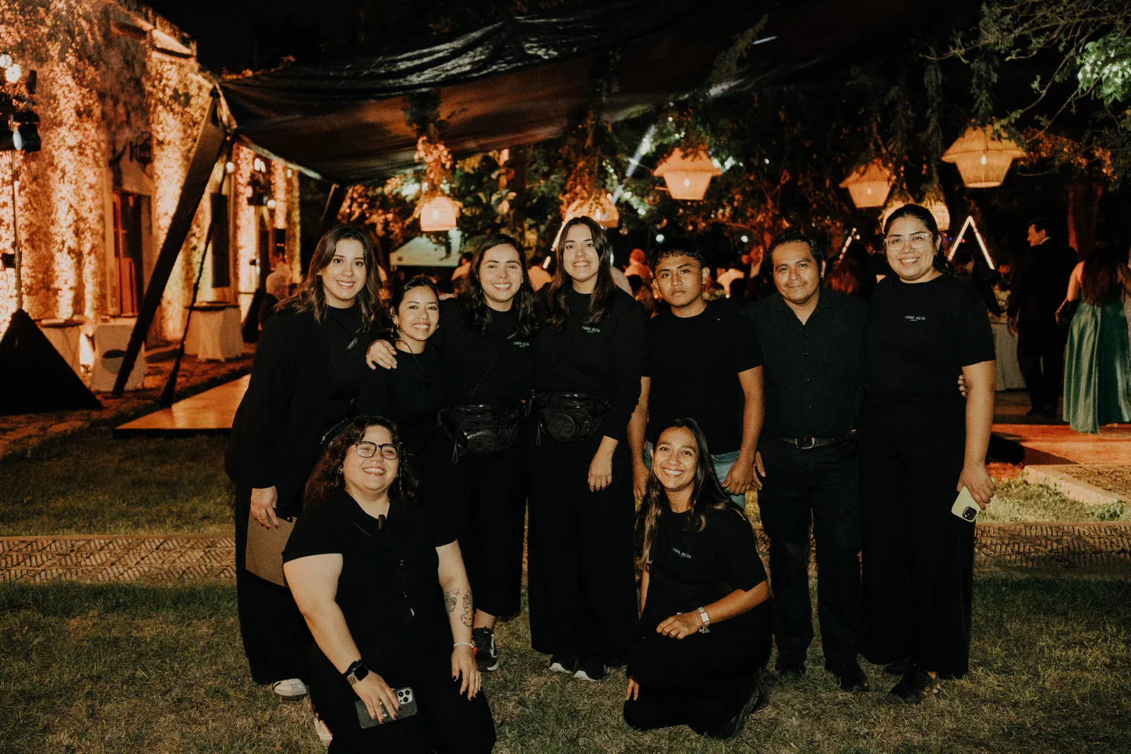 Un grupo de nueve personas, el equipo de Vara Alta, vestidas de negro, posa sonriente en un evento nocturno al aire libre. La cálida iluminación y la vegetación crean un ambiente acogedor.