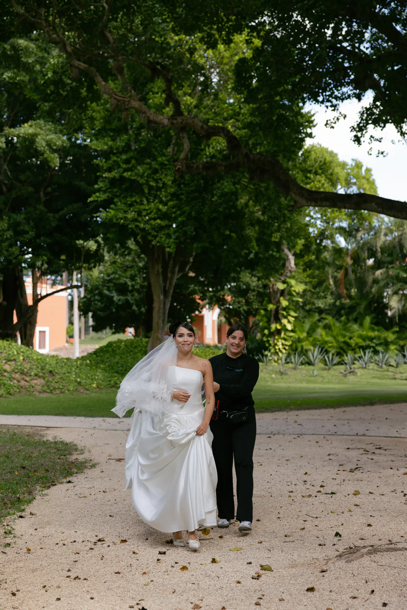 Una novia con un vaporoso vestido blanco y velo camina por un sendero arbolado, acompañada por una persona vestida de negro. El fondo es exuberante y verde.