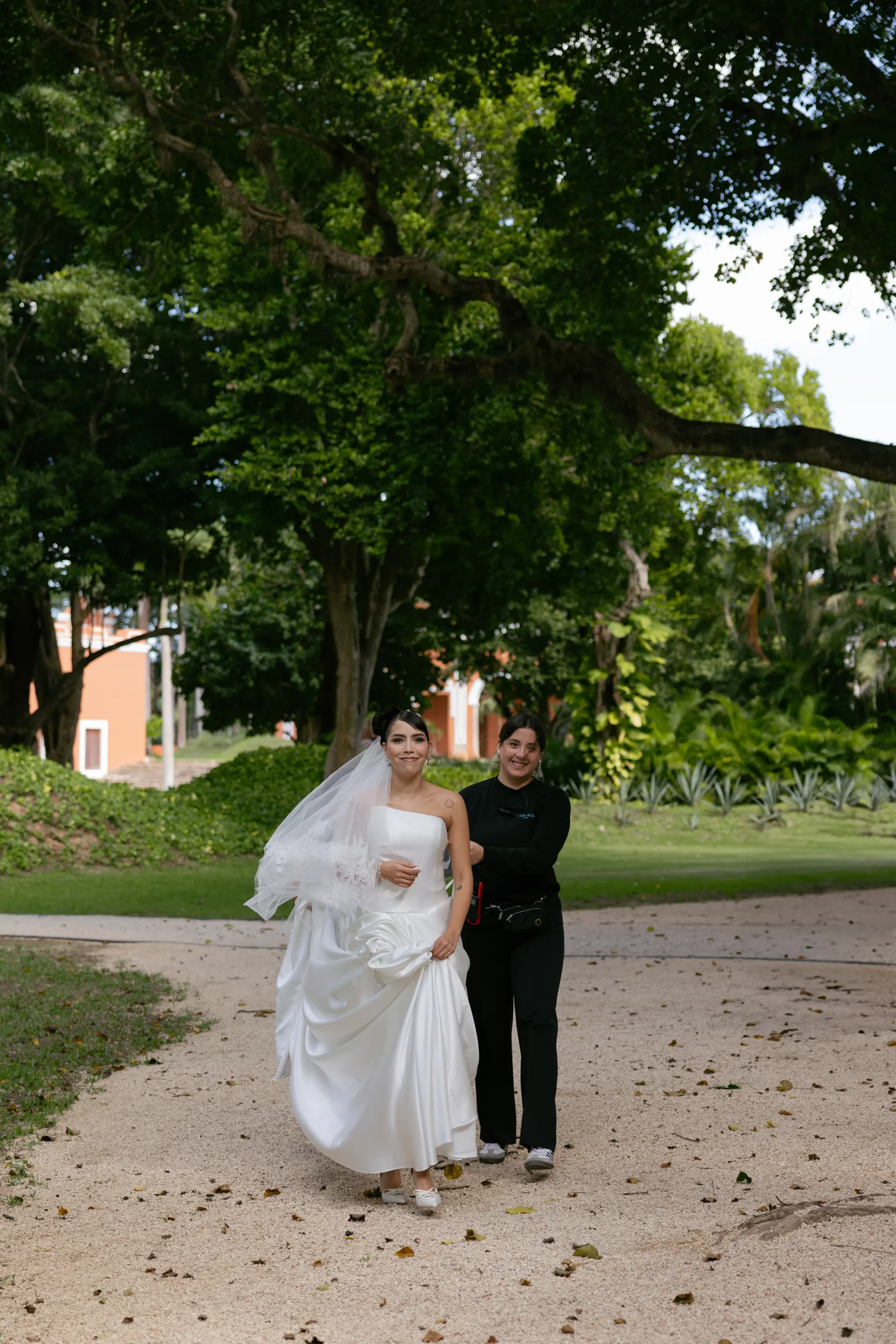 Una novia con un vaporoso vestido blanco y velo camina por un sendero arbolado, acompañada por una persona vestida de negro. El fondo es exuberante y verde.