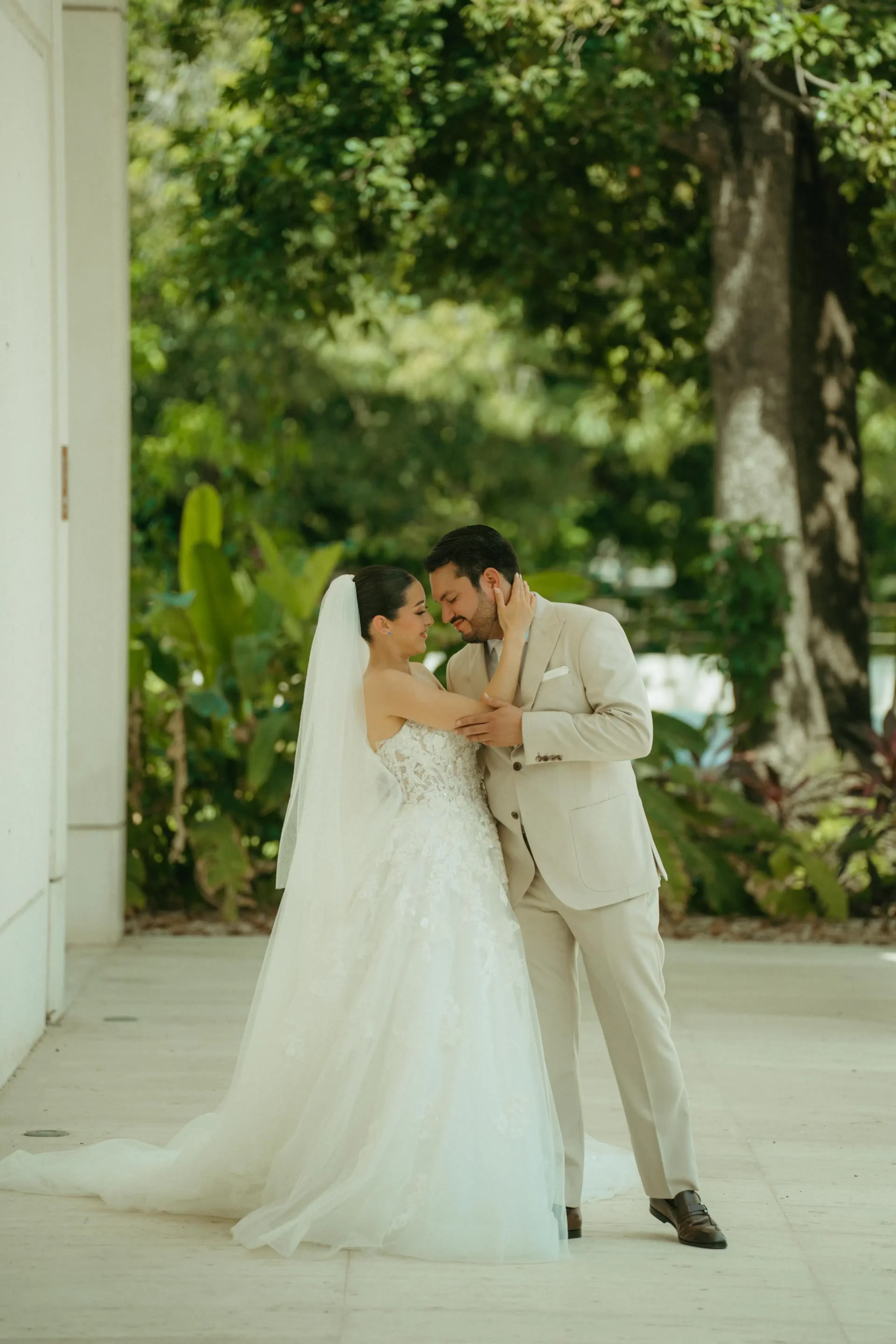 Una novia con vestido blanco y velo abraza a un novio con traje beige, sonriendo con cariño. Están al aire libre, rodeados de exuberante vegetación, lo que transmite alegría y romance.