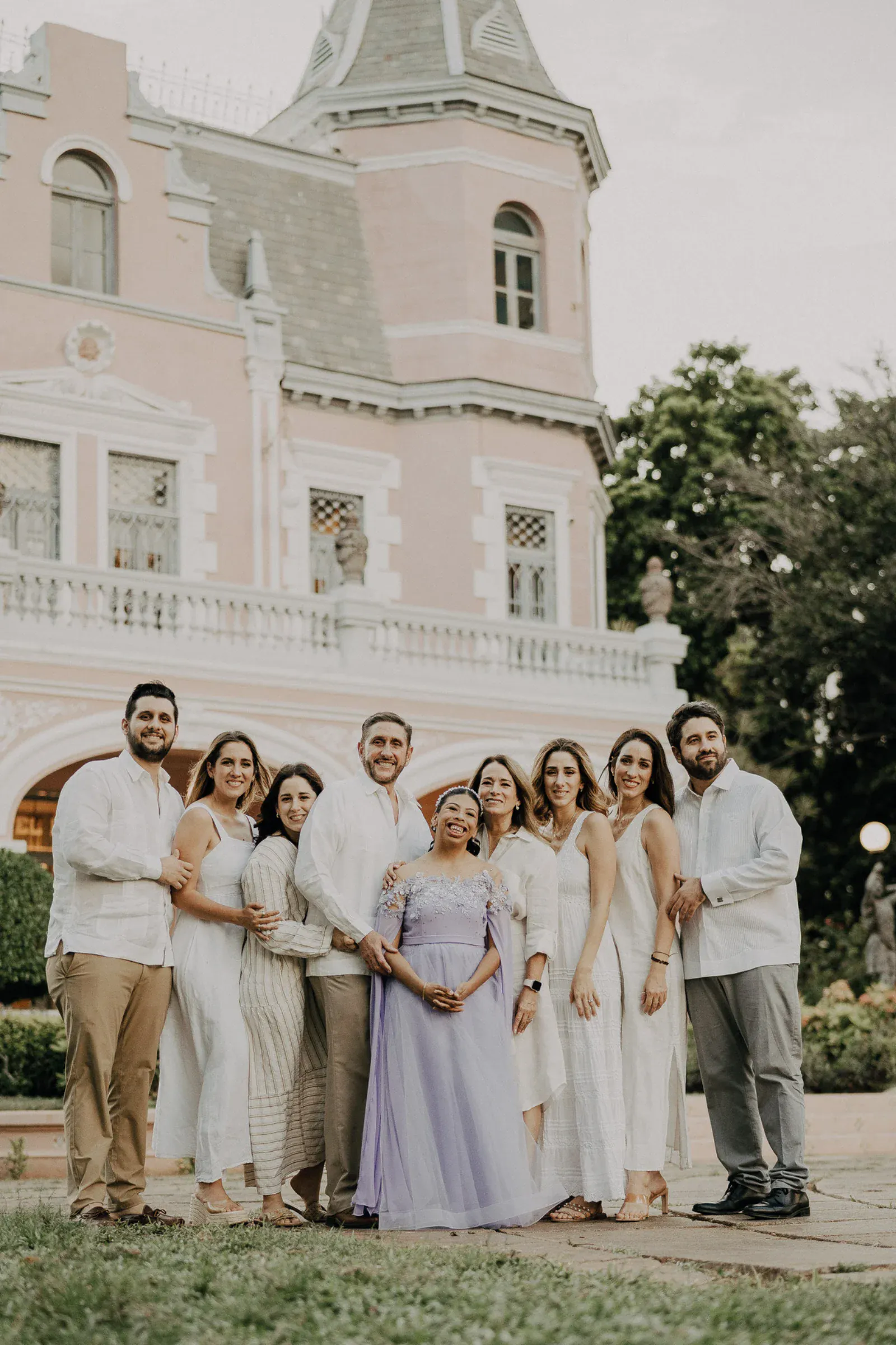 Un grupo de nueve personas, sonrientes y vestidas de blanco, rodea a una mujer con un vestido lavanda. Se encuentran frente a un imponente edificio rosa rodeado de vegetación, transmitiendo alegría y celebración.