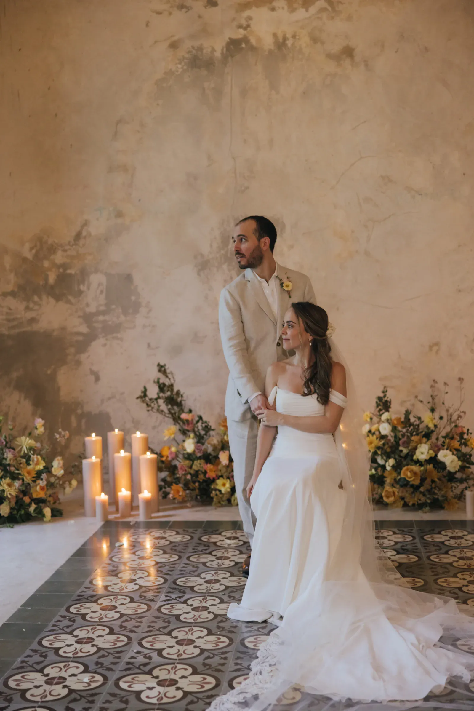 Una novia con un vestido blanco se sienta en un suelo estampado, mirando a su novio con un traje claro. Velas y flores adornan la pared rústica y texturizada. Un ambiente romántico.