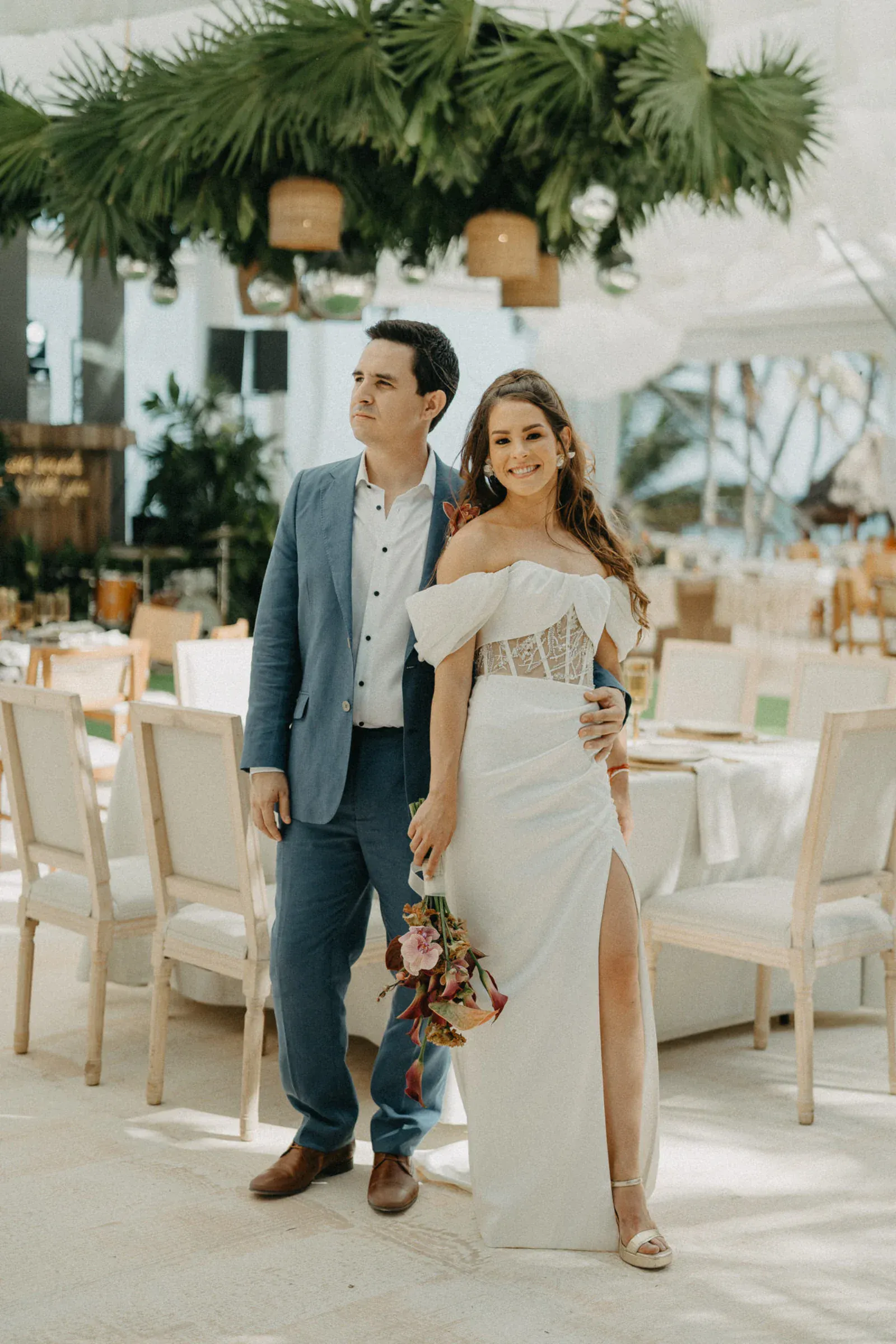 Una pareja en una boda, sonriendo cálidamente. La mujer, con un vestido blanco con una abertura, sostiene flores; el hombre, con un traje azul. Elegante decoración exterior con vegetación.