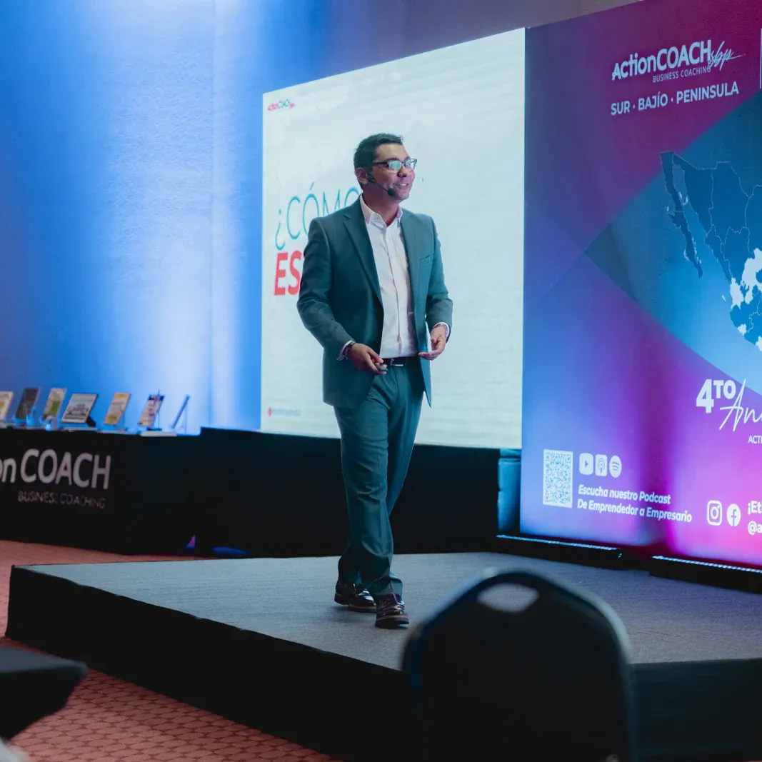 Una persona de traje presenta en el escenario de una conferencia llamada "ActionCOACH", con fondos vibrantes y un mapa. El ambiente es profesional y atractivo.
