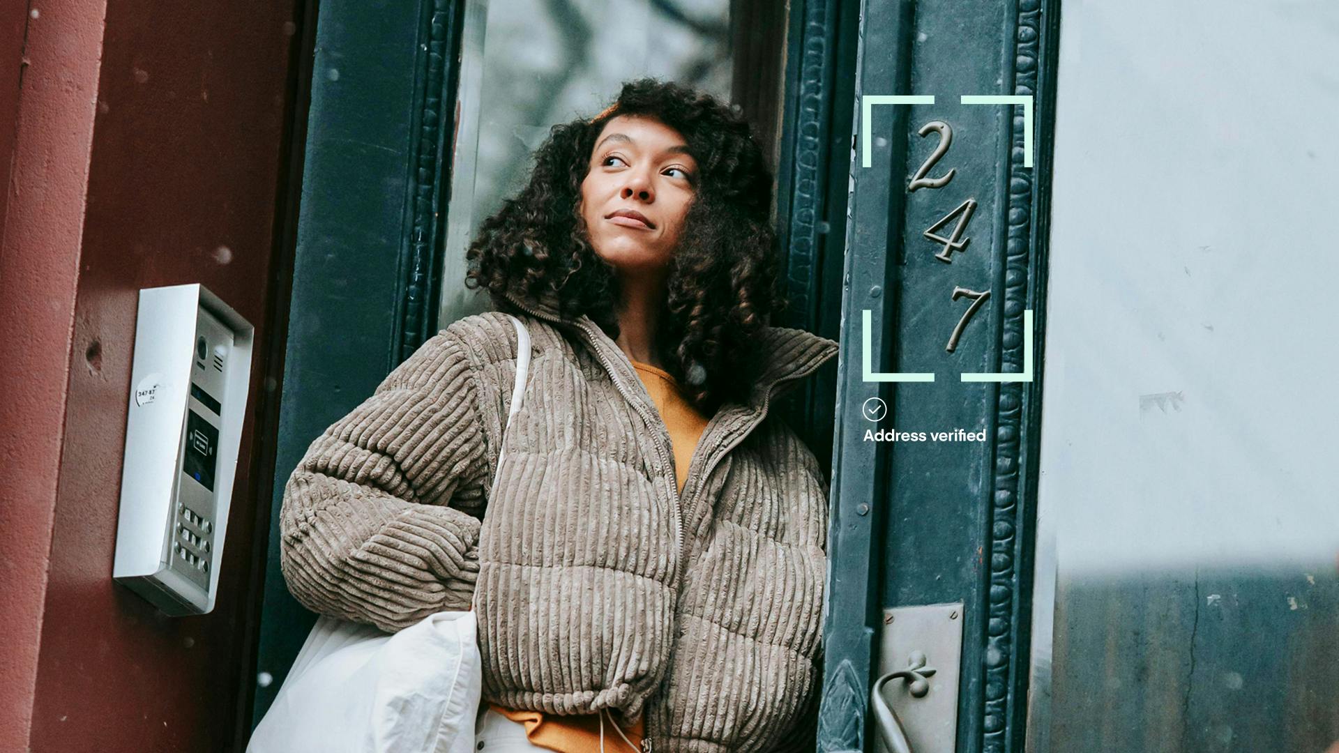 A young woman with curly hair, wearing a brown corduroy puffer jacket, stands in a doorway looking upwards, with a "247" digital overlay and "Address verified" text superimposed to her right.