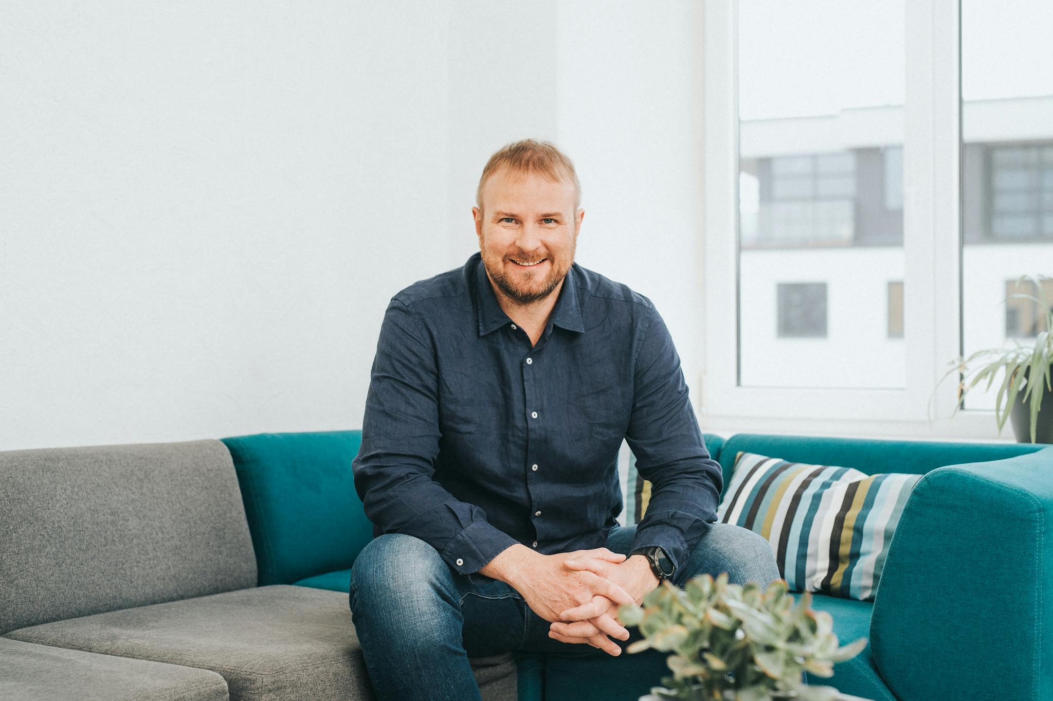 A man sat down smiling on a sofa in a blue shirt