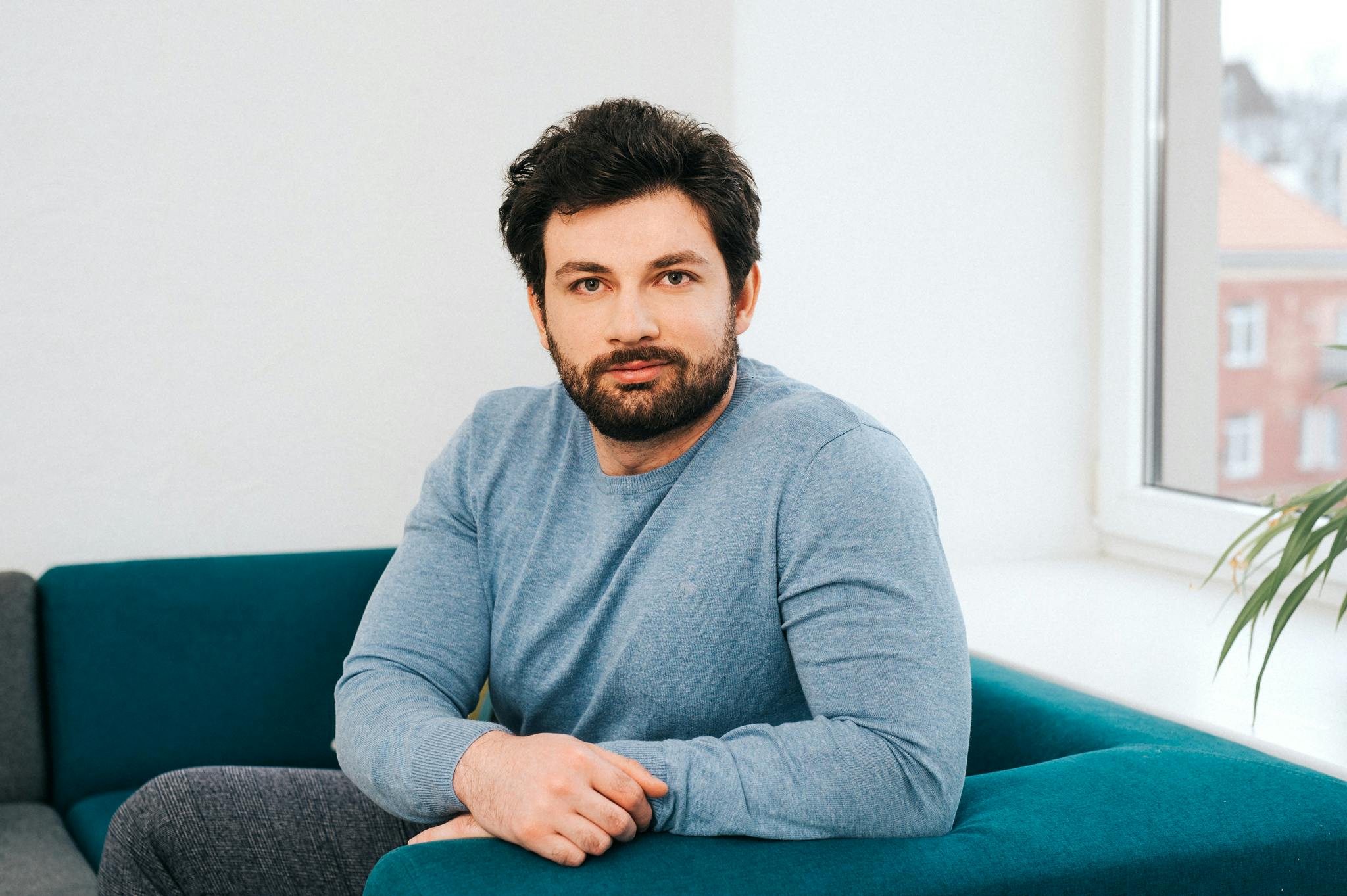 A dark haired man with a beard sat on a sofa smiling