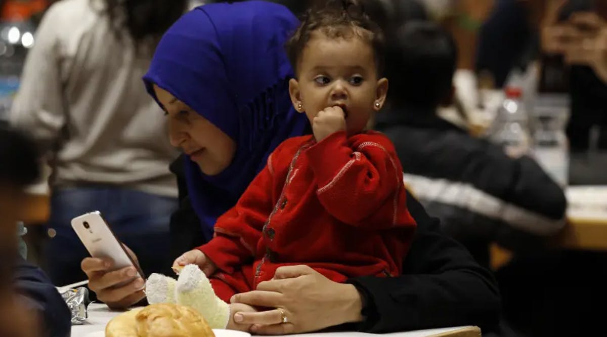A migrant woman from Syria checks her mobile device as she eats with her daughter