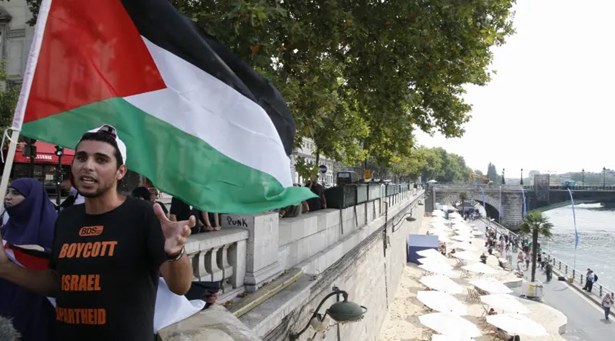 A man wearing a T-shirt with the message, "Boycott Israel Apartheid" holds a Palestinian flag during a protest action on a bridge overlooking umbrellas placed along the artificial beach along the "Paris Plages"