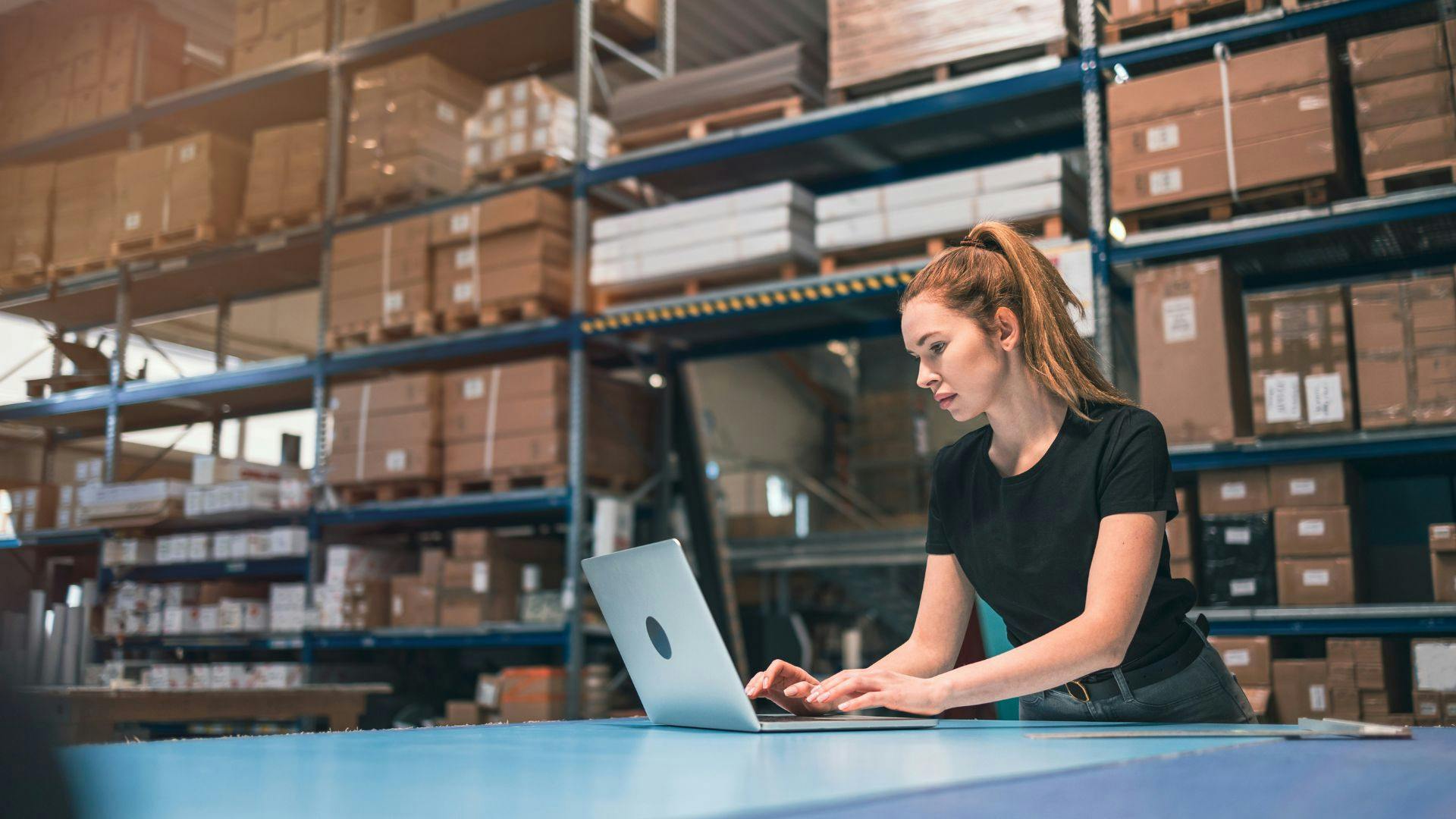 Image of a woman in a logistics warehouse, standing and focused, typing on a laptop. Surrounding her, numerous packages and boxes on shelves fill the background, creating an industrious atmosphere.