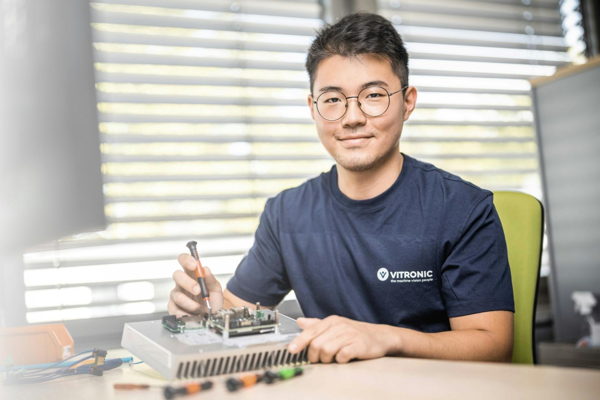 Young trainee at VITRONIC sitting at a desk working on hardware components of an image processing system.