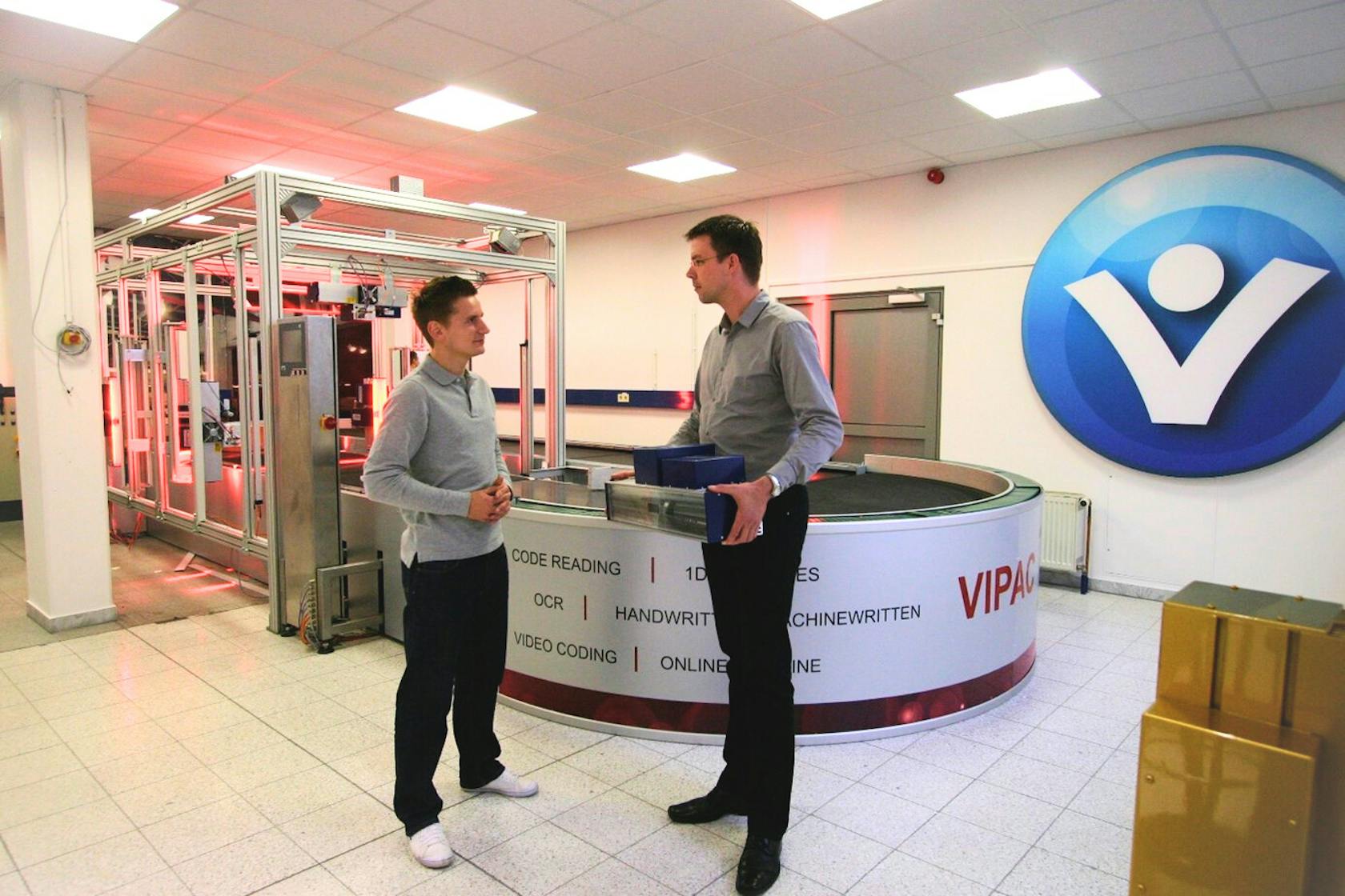 Zwei Männer sprechen miteinander vor einer Logistik-Sortieranlage in einem hellen Raum. 