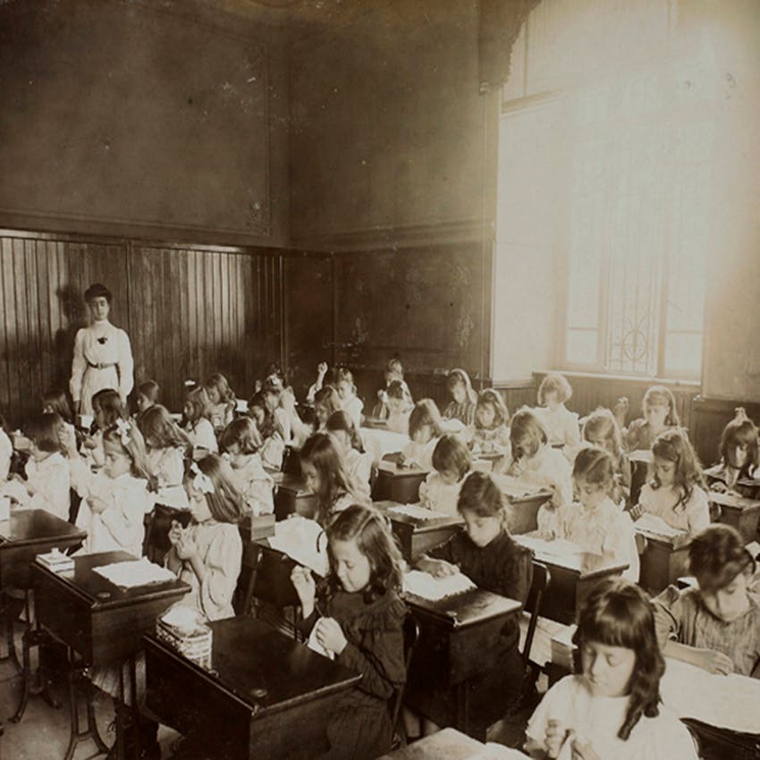 Foto antiga em plano aberto, em preto e branco, de uma sala de aulas só com meninas. Elas estão sentadas em antigas carteiras de madeira e a sala é iluminada por uma janela grande em formato de arco.