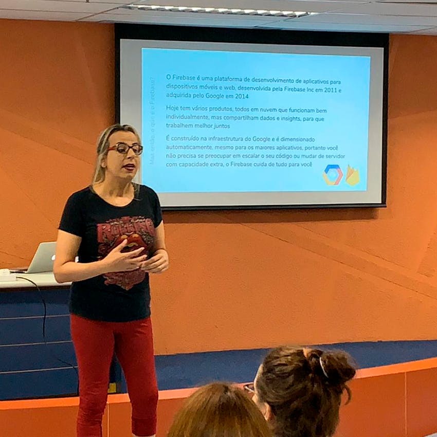 Foto em plano fechado de um pequeno auditório. À frente, há o telão e uma mulher branca, de cabelos longos e loiros, óculos, camiseta preta e calça vermelha, fazendo uma apresentação.